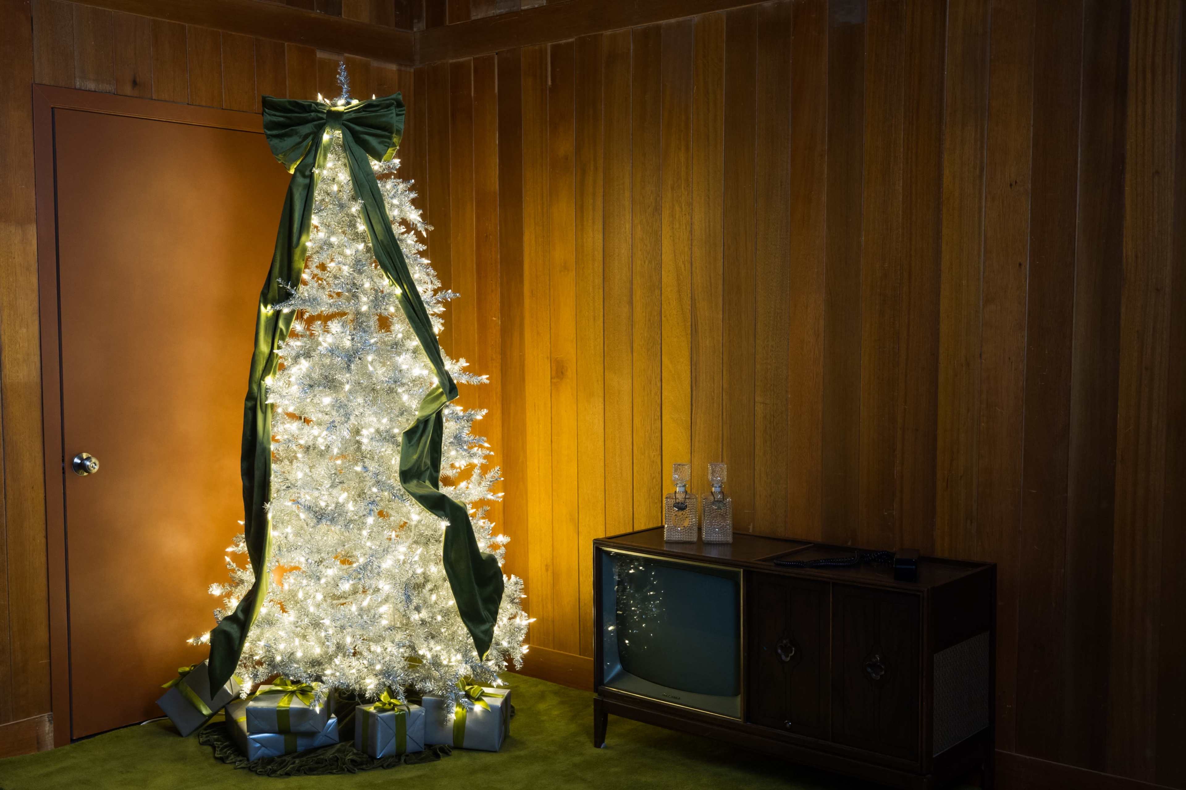 A decorated Christmas tree with white lights and green ribbons stands beside a wooden wall, next to a vintage television and wrapped gifts.