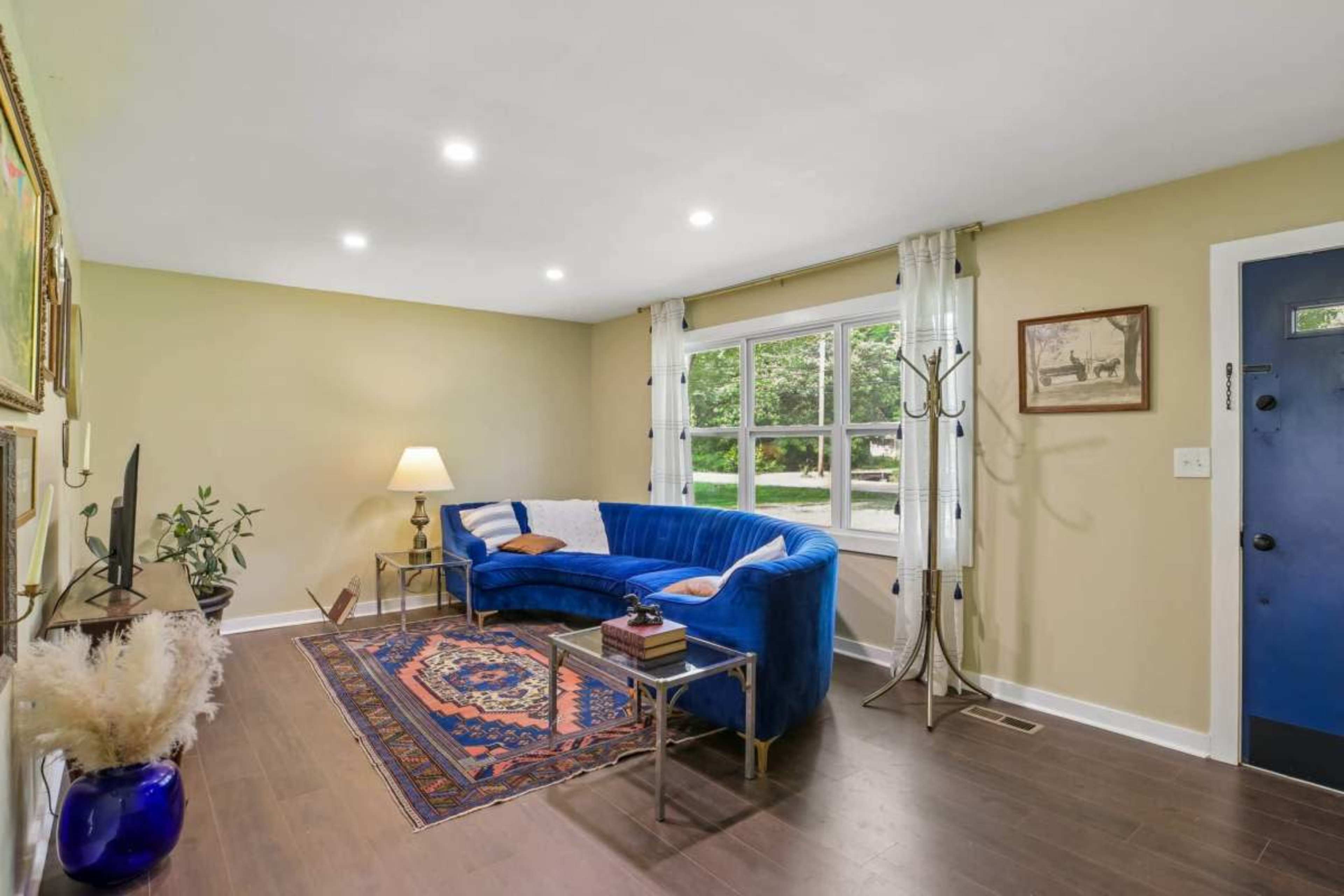 The image shows a living room featuring a blue sectional sofa, a lamp, and a window that overlooks a green outdoor area.