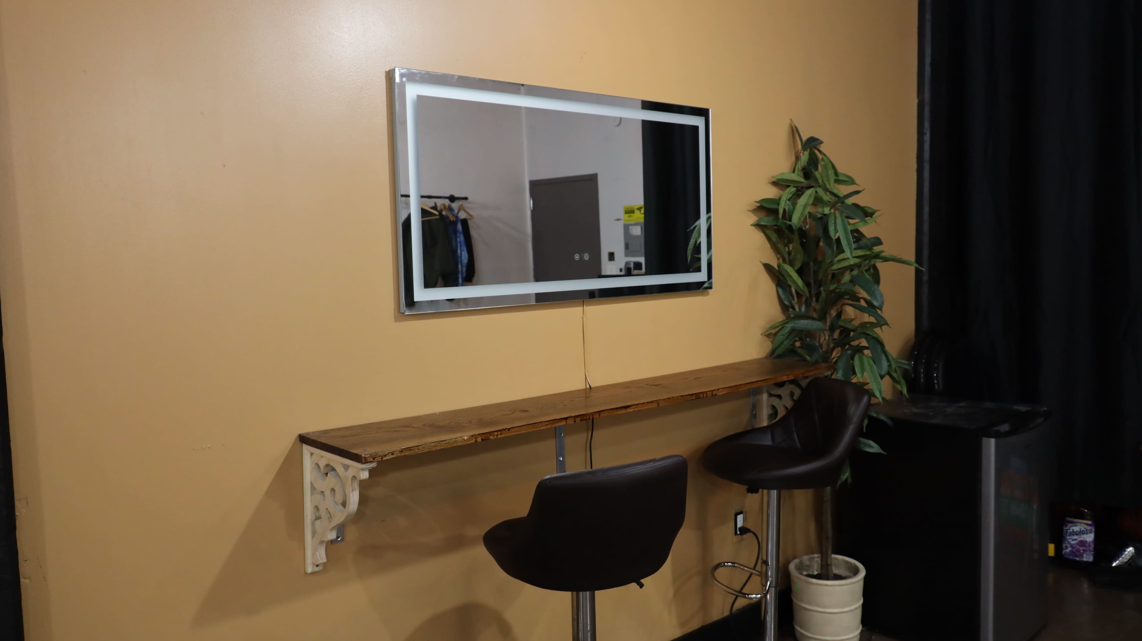 A wall-mounted mirror is positioned above a narrow wooden shelf with two black stools beside a potted plant on a beige wall.