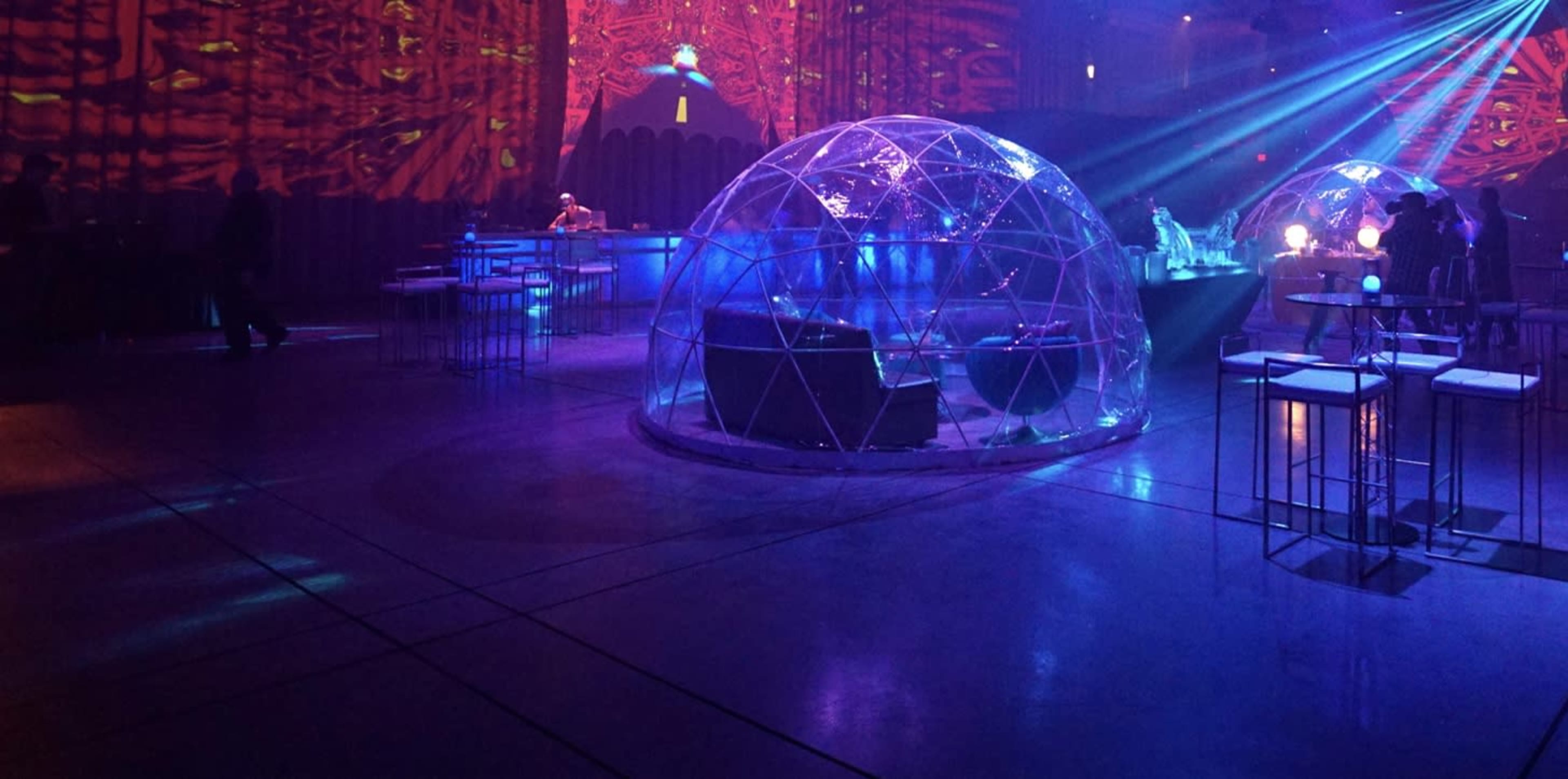 The image shows a dimly lit event space featuring illuminated geodesic domes, surrounded by high tables and a colorful light display on the walls.