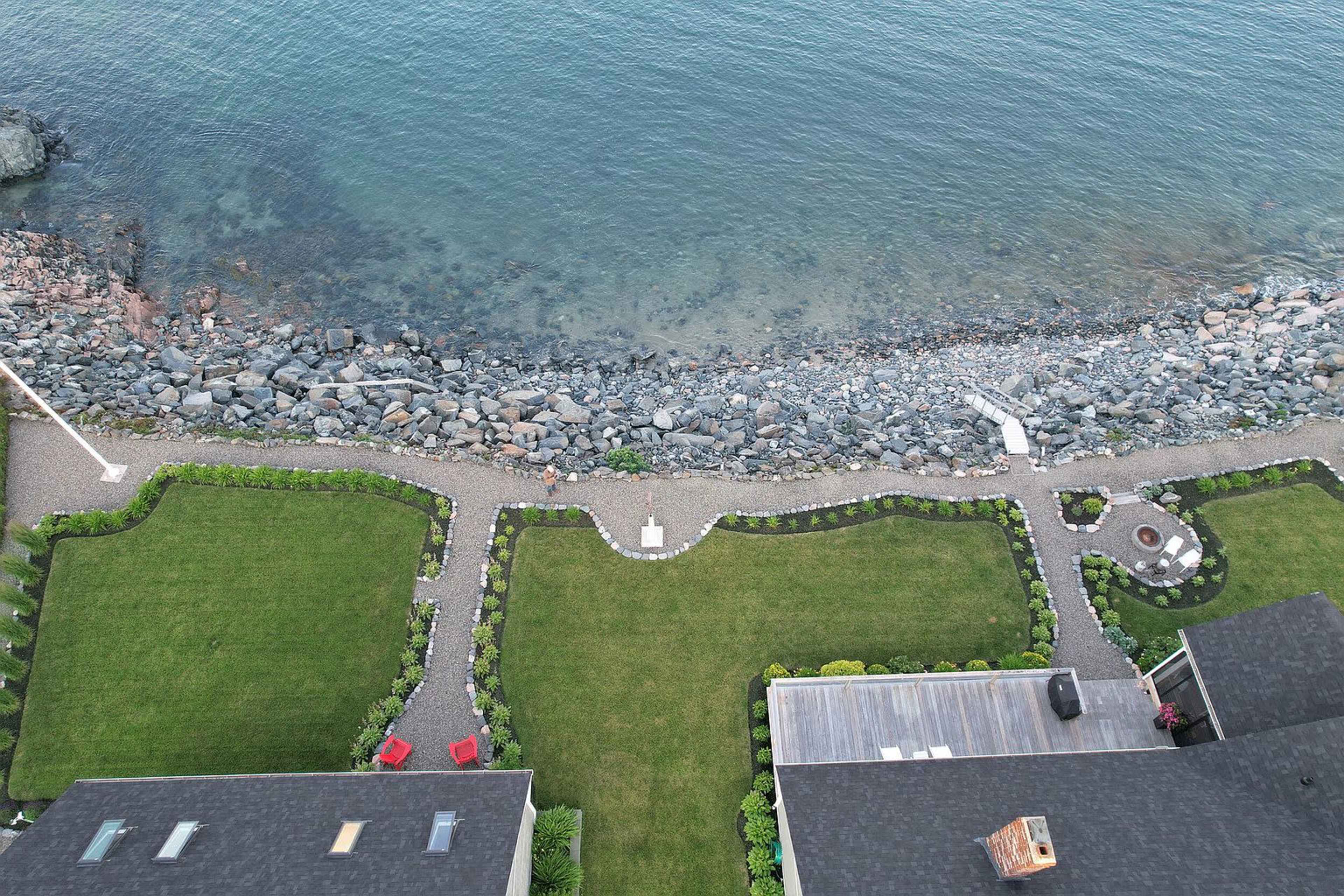 The image shows a coastal property with two neatly landscaped green lawns, bordered by a gravel path and rocky shoreline leading to the water's edge.