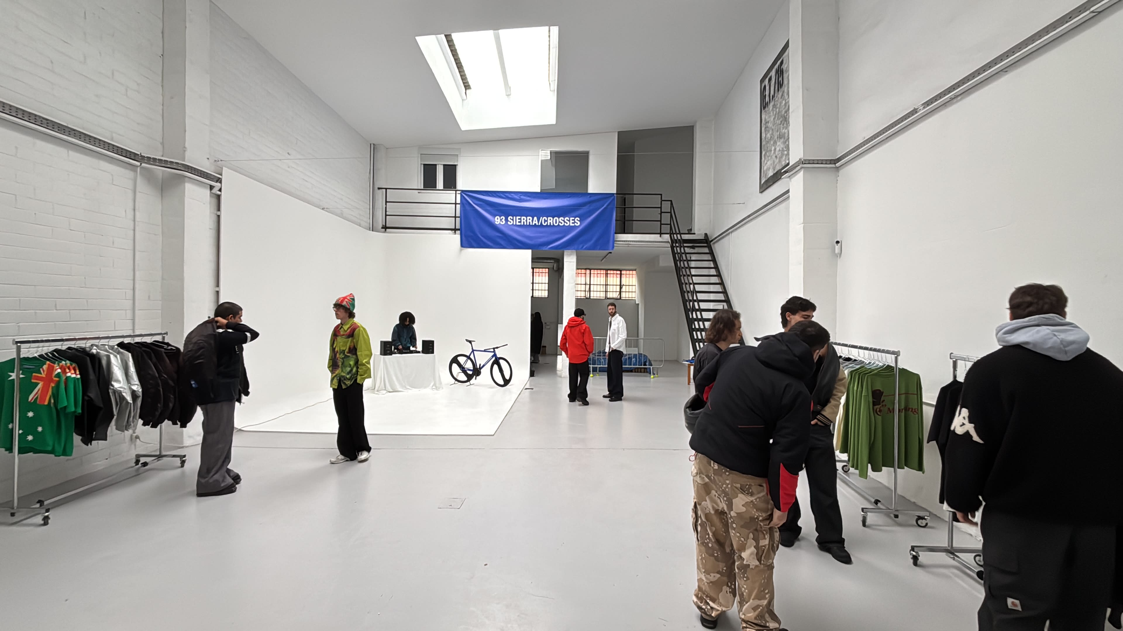 The image shows a modern, spacious interior with a few people browsing clothing racks and a central display area featuring a bicycle and a seated person behind a table.