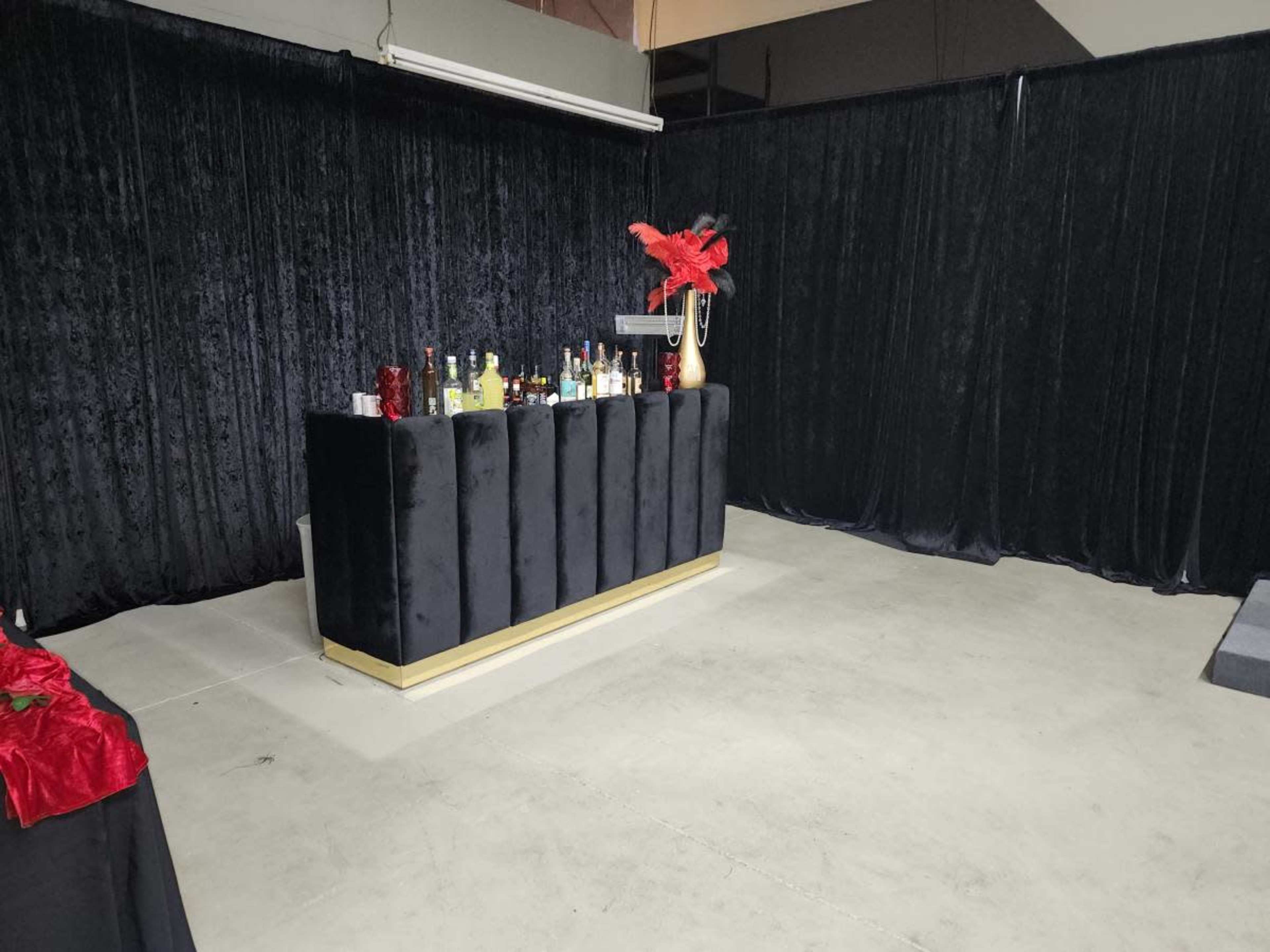 The image shows a bar counter covered with an assortment of liquor bottles, set against a backdrop of black curtains in a spacious room with a concrete floor.