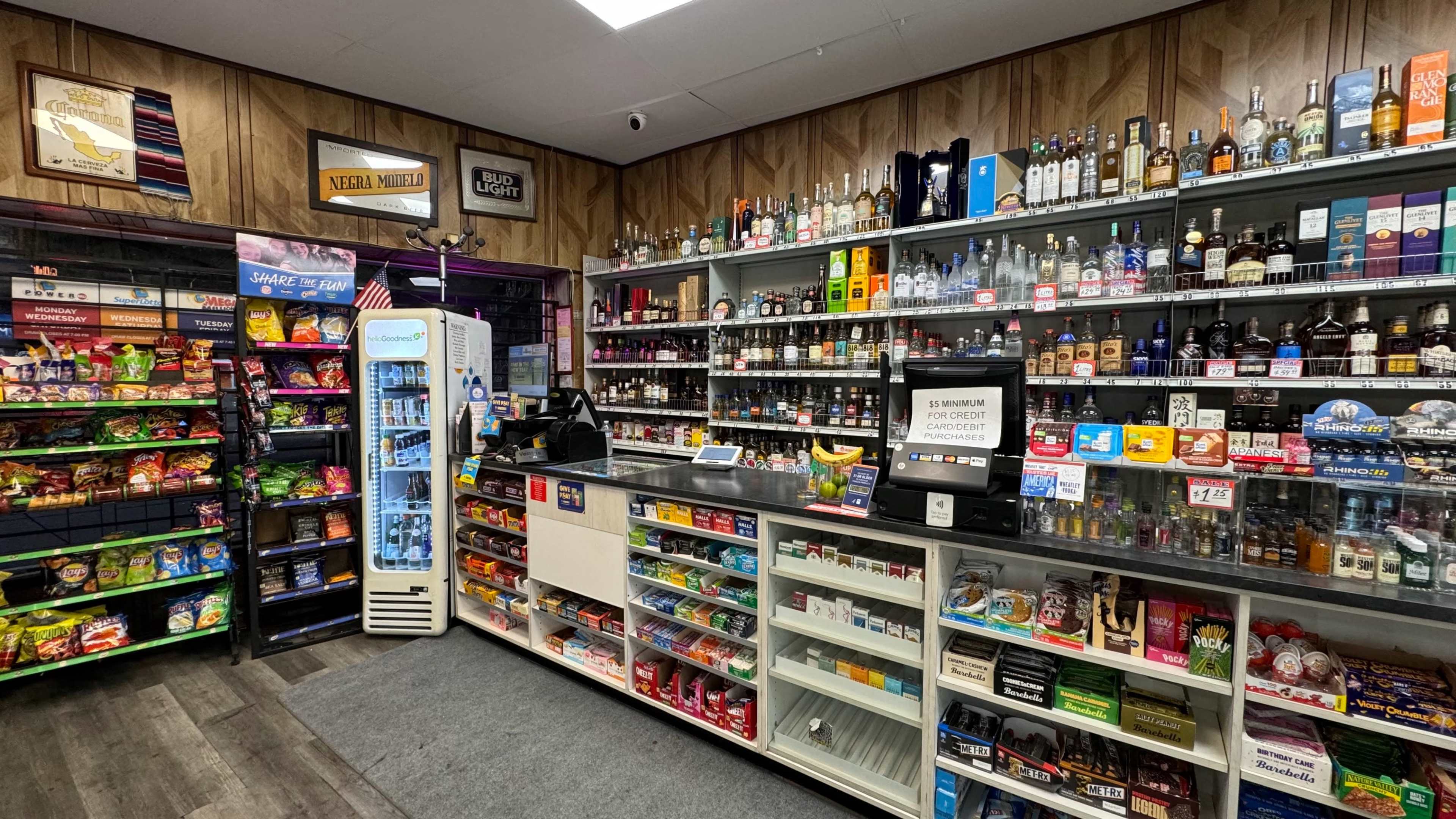 The image shows the interior of a convenience store with shelves of snacks, beverages, and assorted alcoholic drinks displayed behind a cash register.