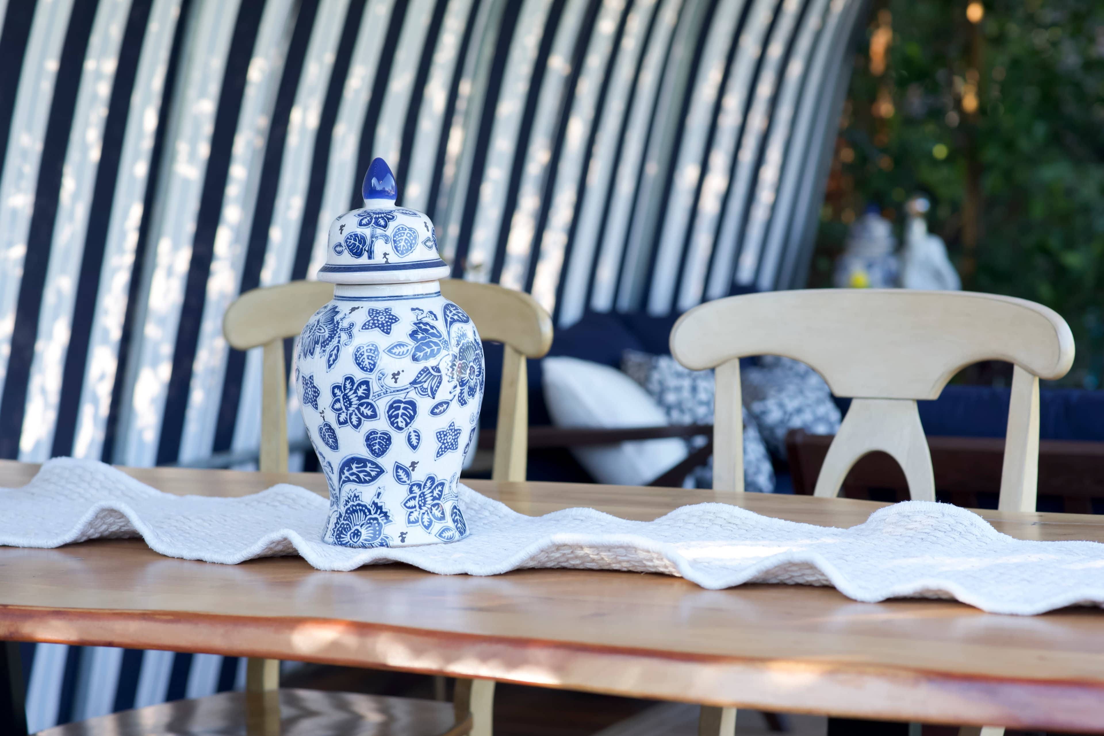 A blue and white patterned ceramic jar sits on a wooden table with a textured white runner, while striped walls are visible in the background.
