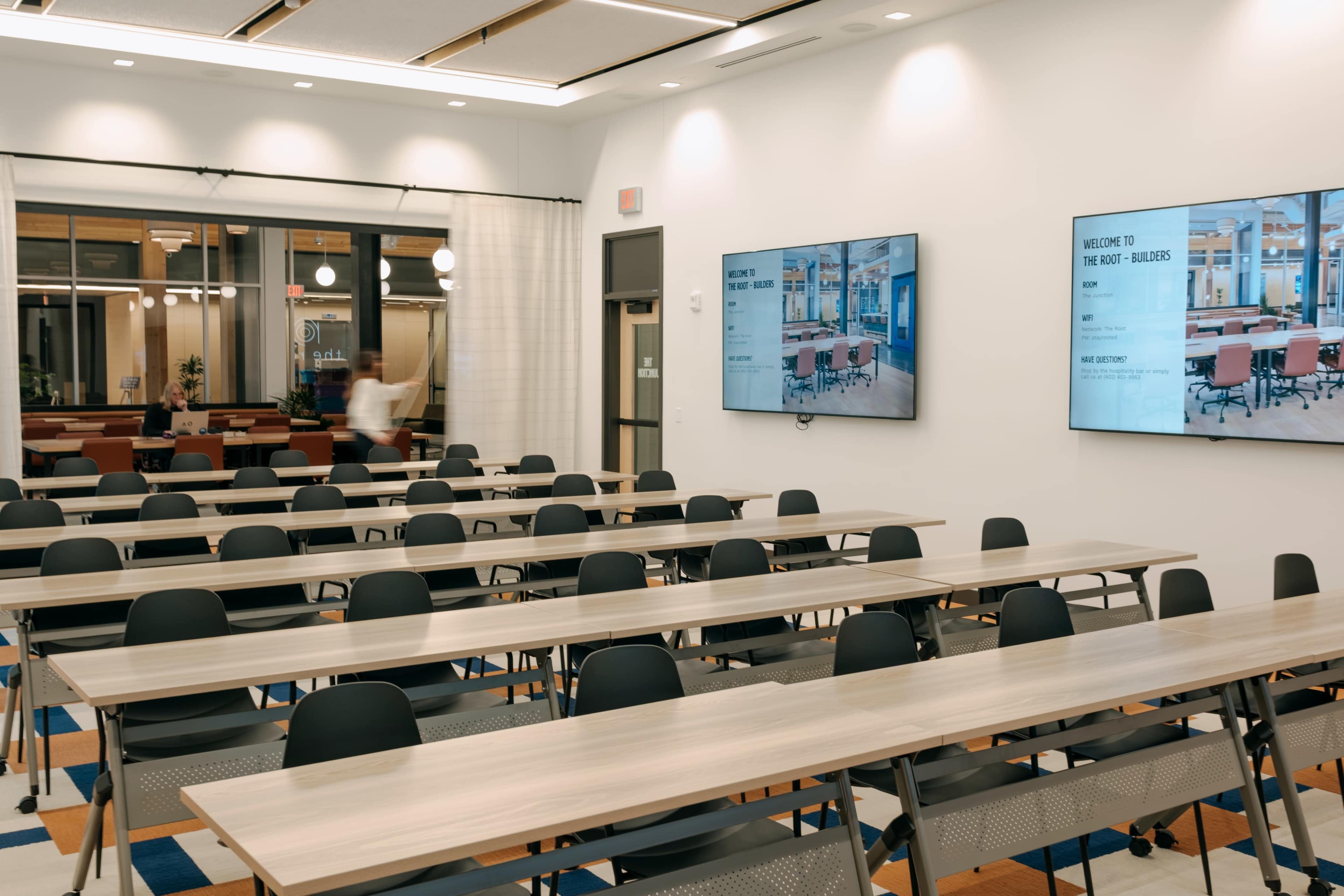 The image shows a modern classroom or training room featuring multiple rows of tables and chairs, with two large screens displaying information on the walls.