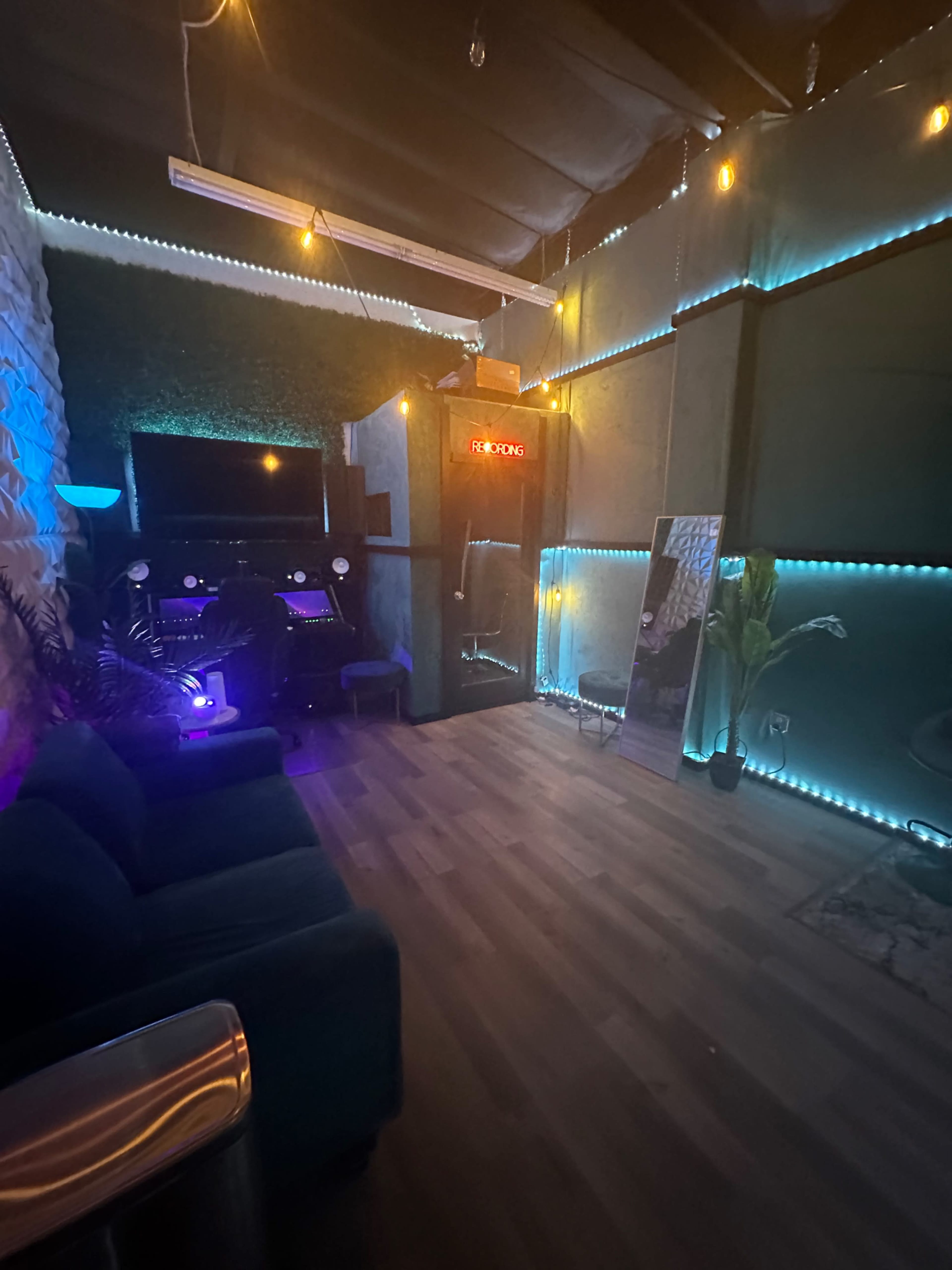 A cozy interior space with blue and green lighting, featuring a seating area with a couch, a music workstation, and decorative plants.