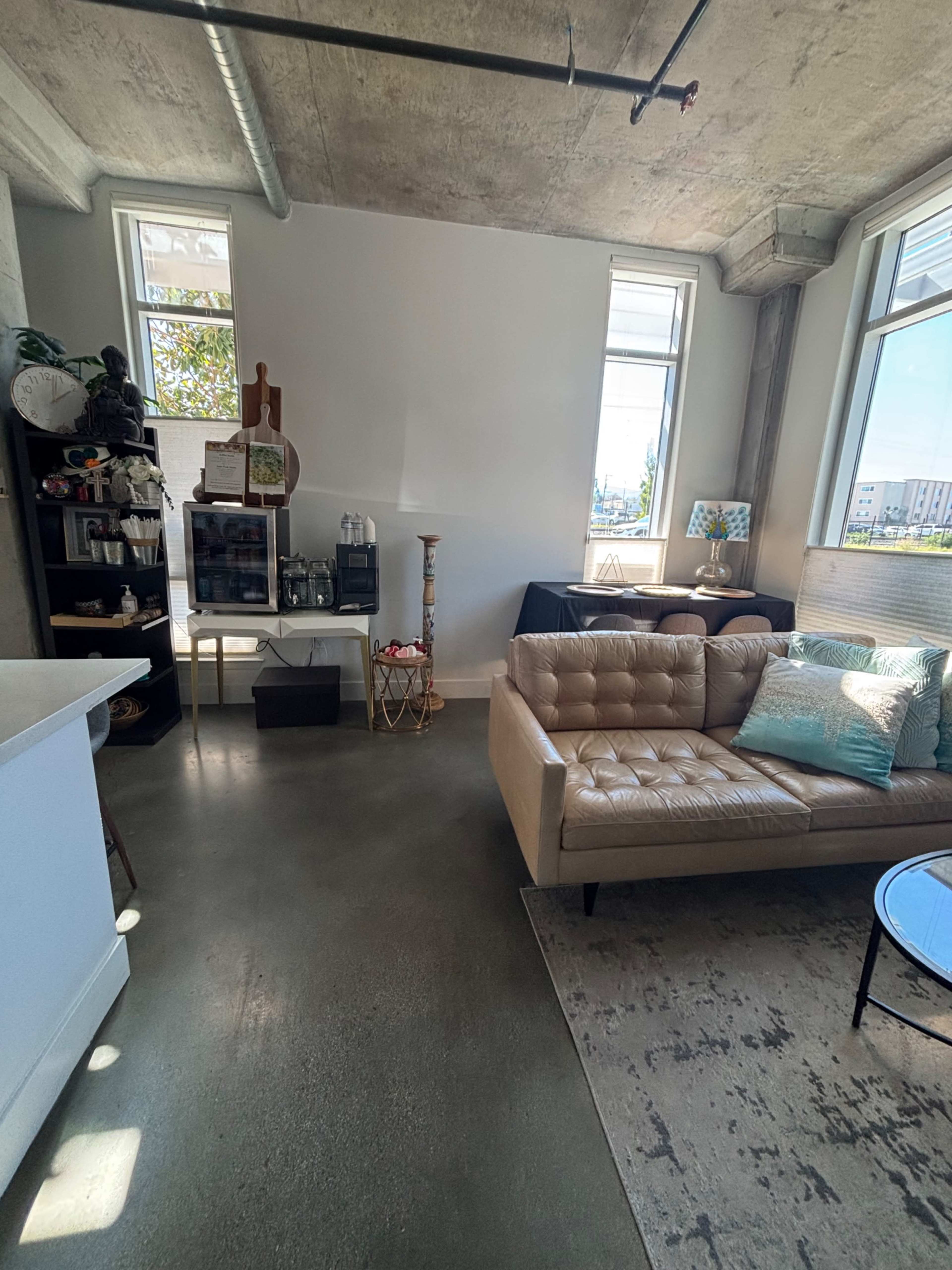 The image shows a modern, industrial-style living space featuring a beige couch, a glass coffee table, and various decor items on shelves and tables, with large windows allowing natural light to enter.