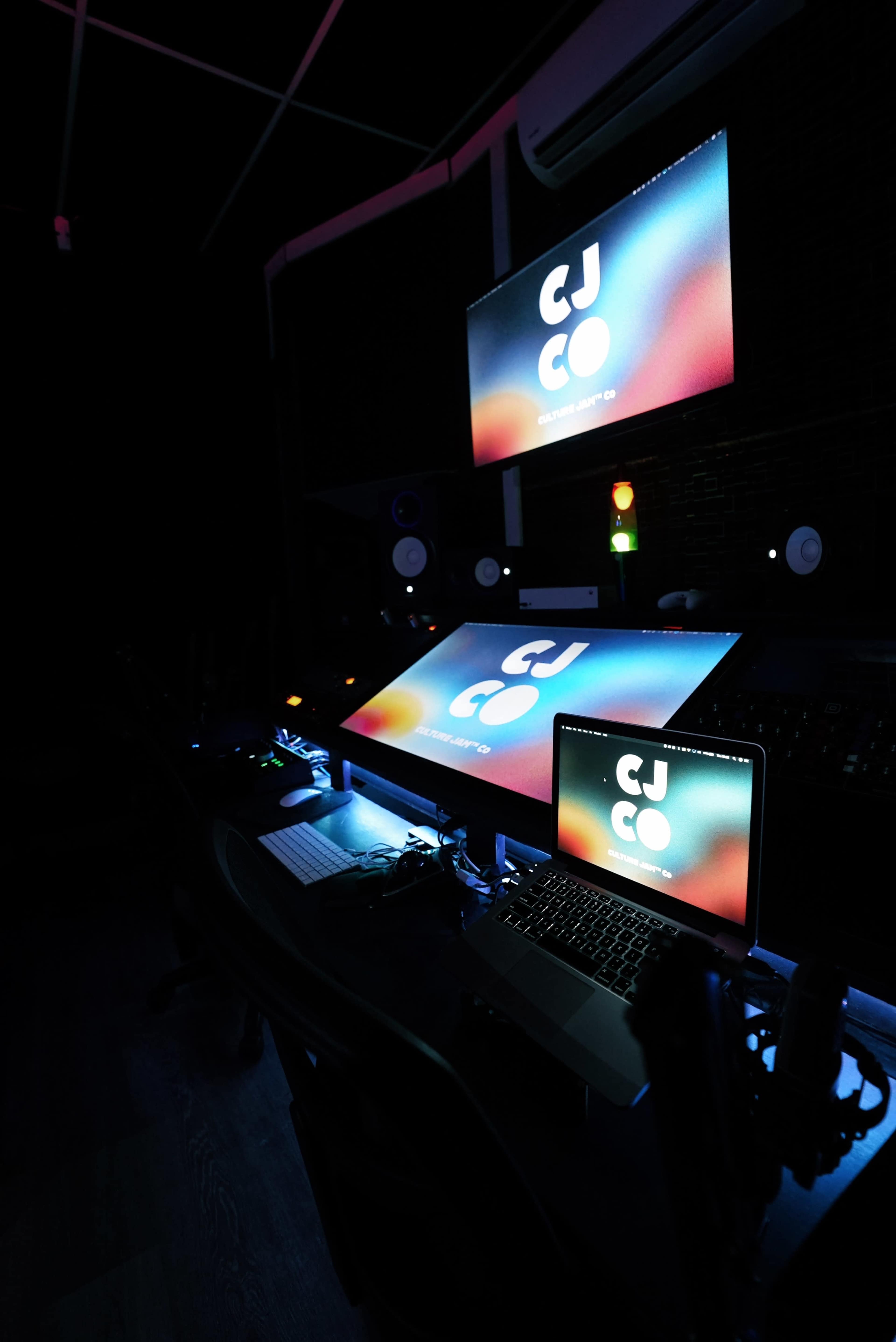 The image shows a dimly lit audio or video production studio with multiple computer screens displaying the text "CJ CO" and various equipment arranged on the desk.
