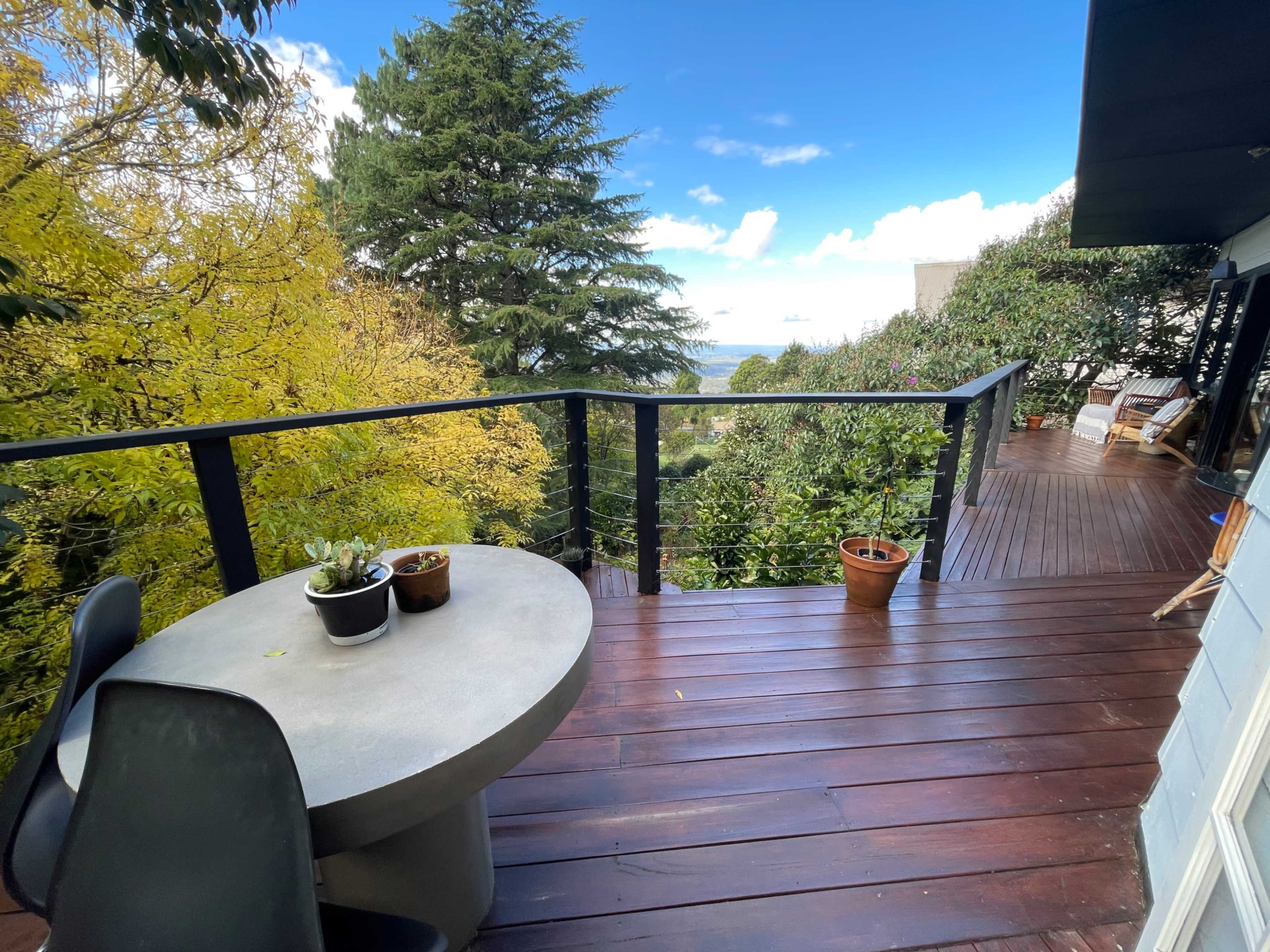 A wooden deck features a round table and potted plants with views of trees and a clear sky in the background.