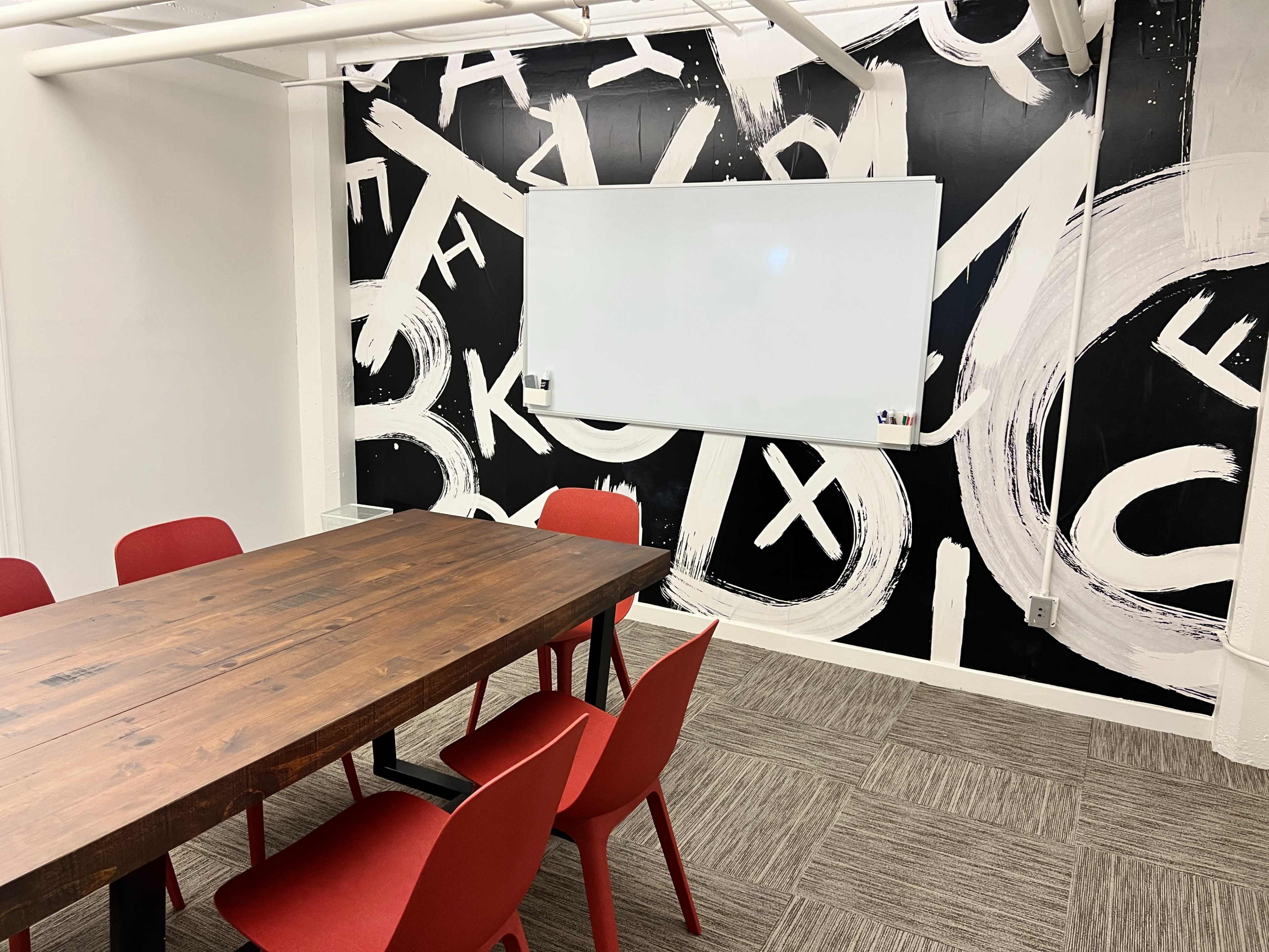 A conference room features a wooden table surrounded by red chairs, with a whiteboard mounted on a wall painted with abstract black and white letters.