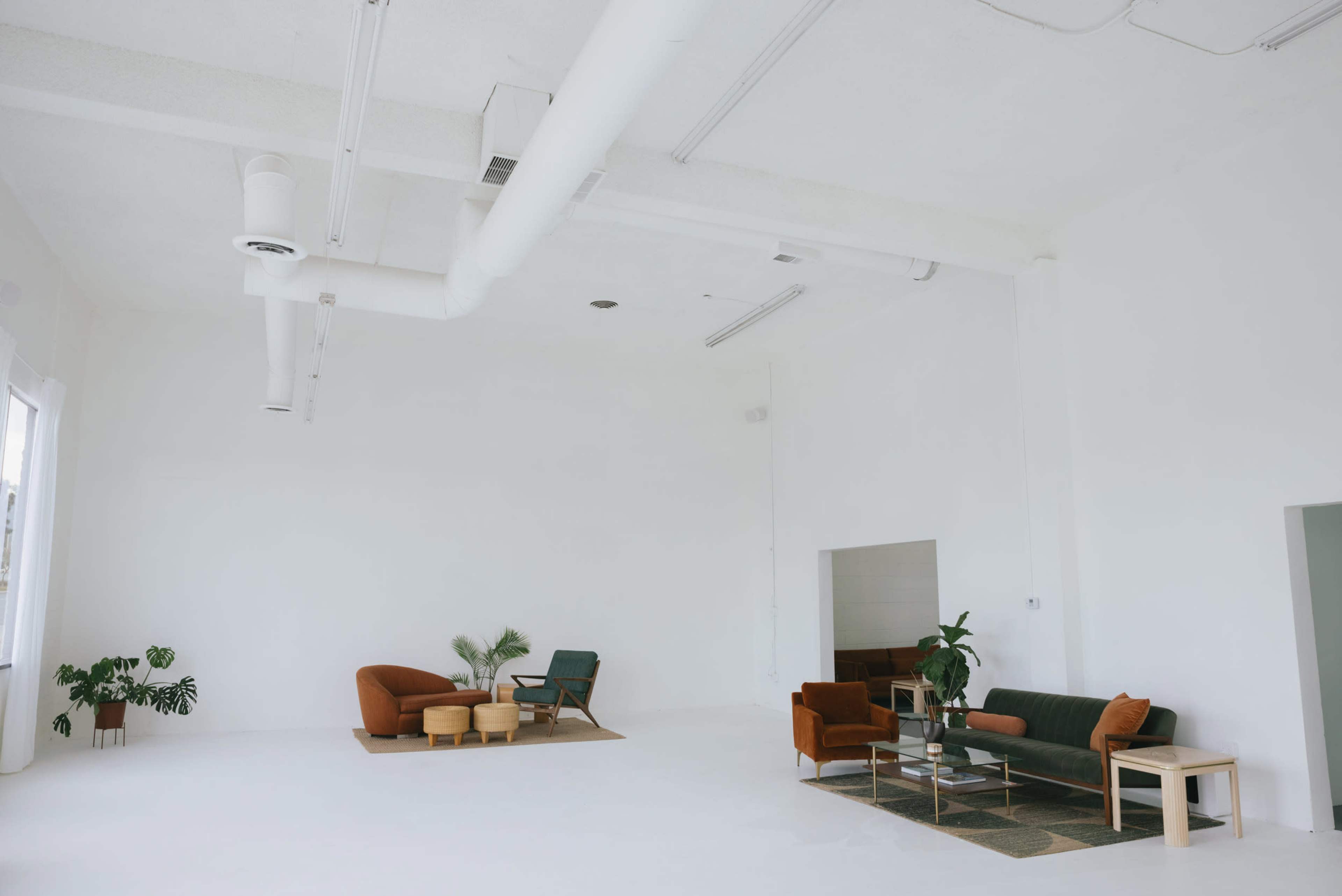 The image shows a spacious, minimalist interior with white walls and a few distinct seating areas arranged with furniture and plants.