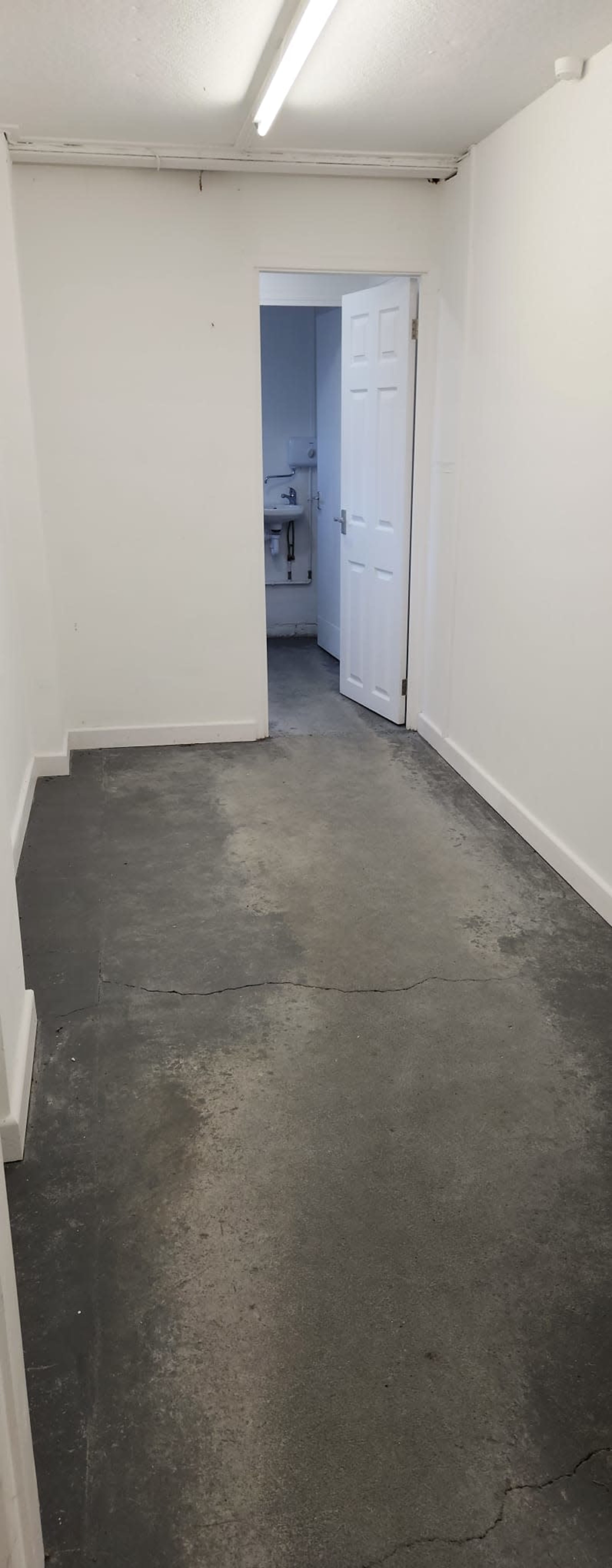 The image shows a blank, empty room with gray concrete flooring and white walls, featuring an open door at the end leading to another room.