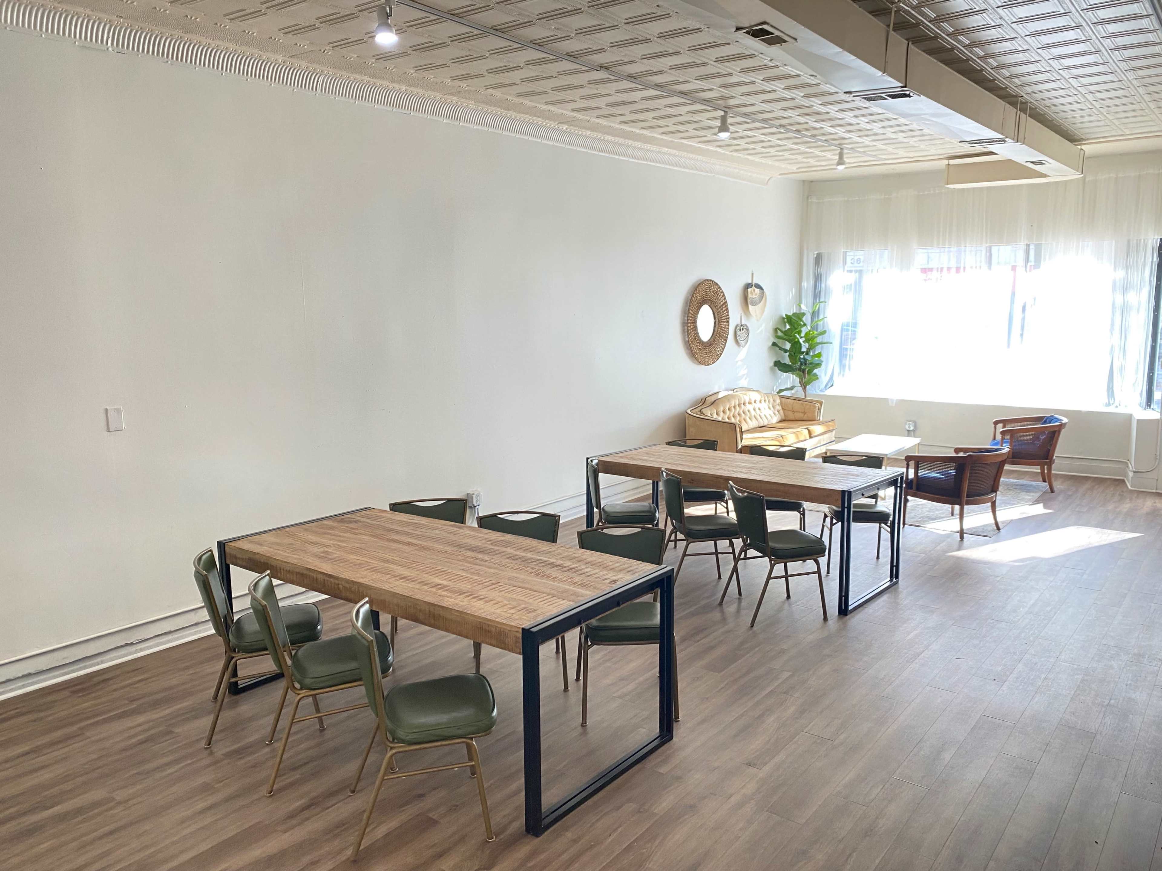 A spacious, well-lit room with several wooden tables and chairs arranged, along with a sofa and a large window letting in natural light.