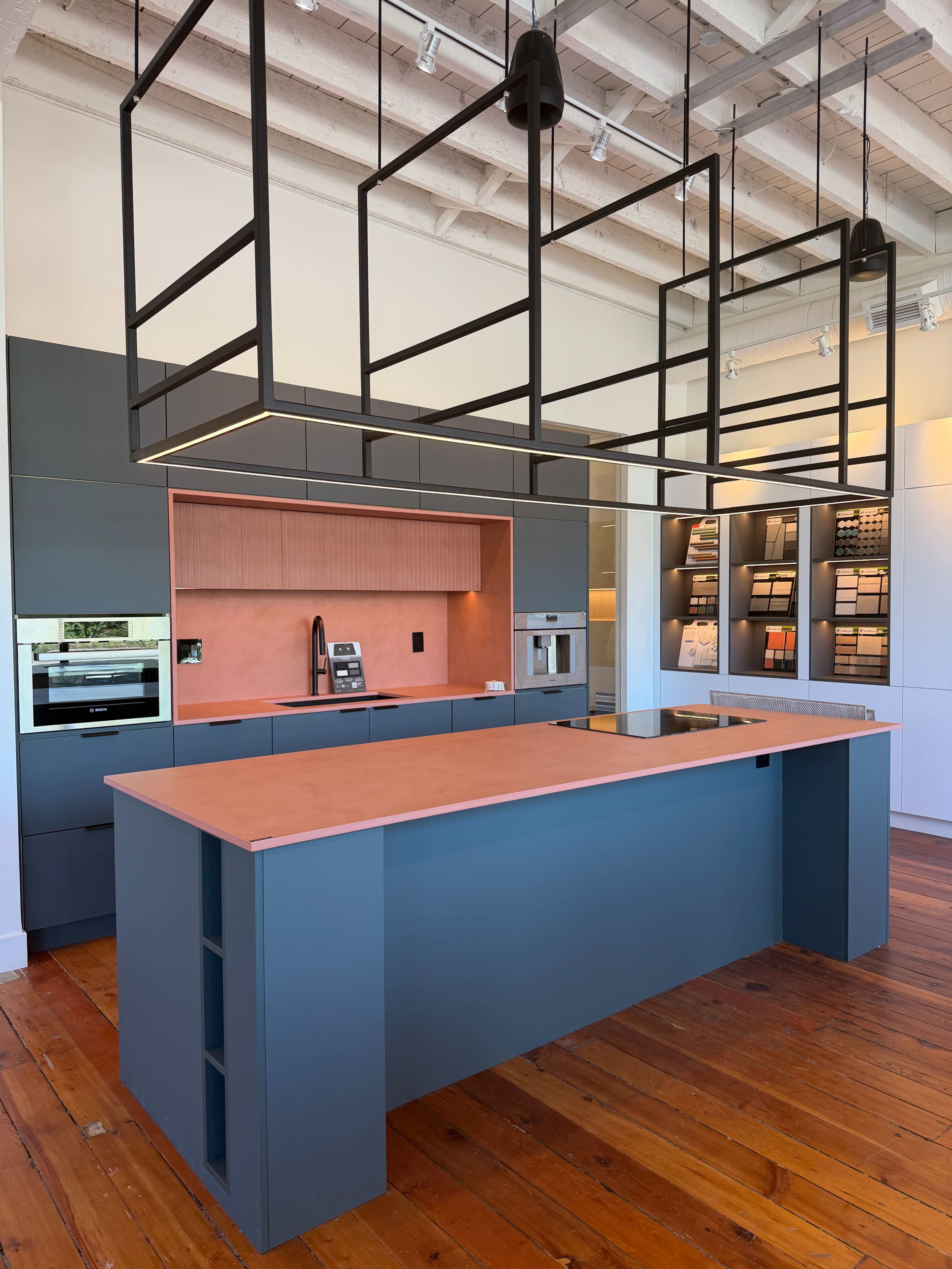 The image shows a modern kitchen with a central island, featuring a copper-colored countertop and dark cabinetry, illuminated by a geometric light fixture above.