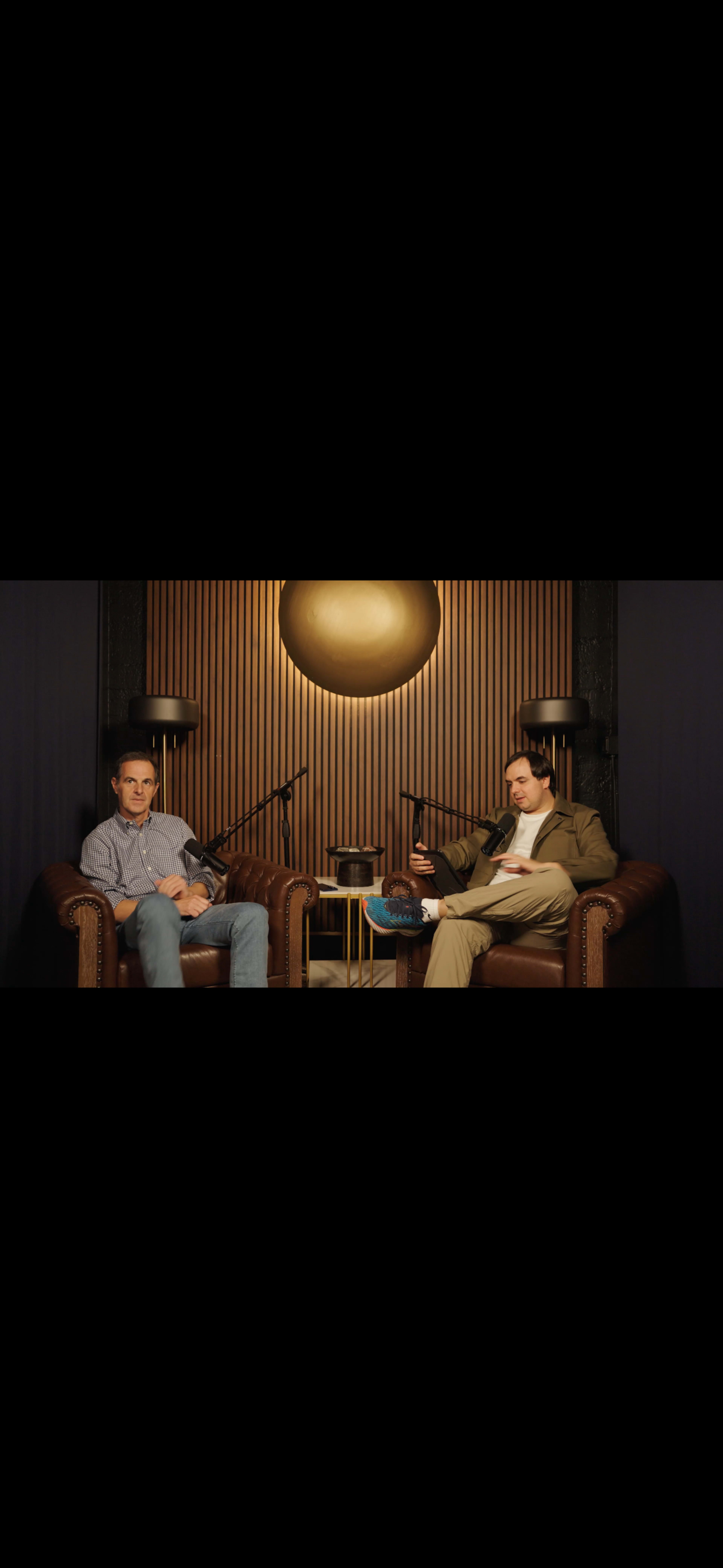 Two men sitting on leather armchairs in a dimly lit studio with a circular light fixture in the background.