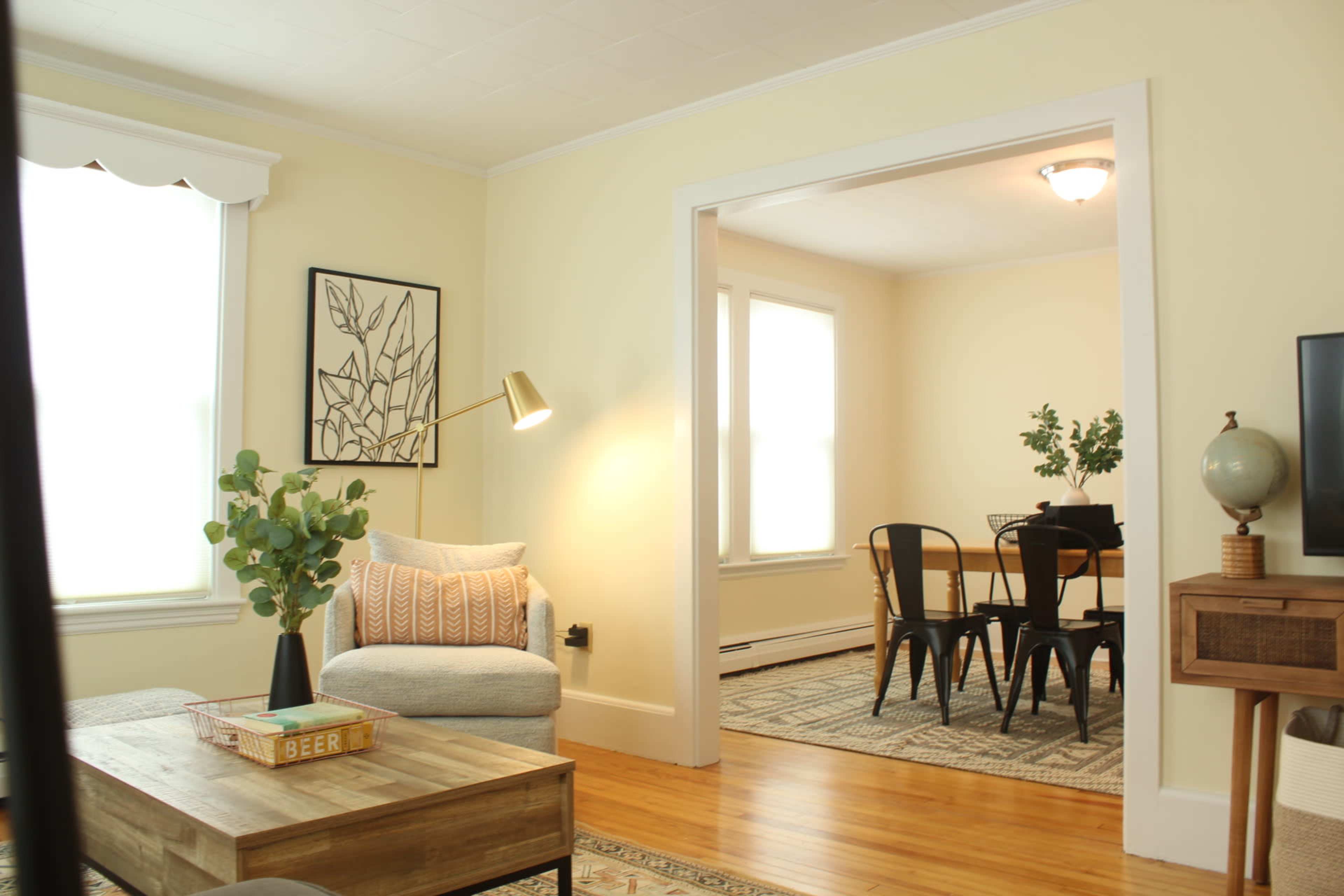 A well-lit living room features a sofa, a wooden coffee table, and a view into an adjacent dining area with a table and chairs.
