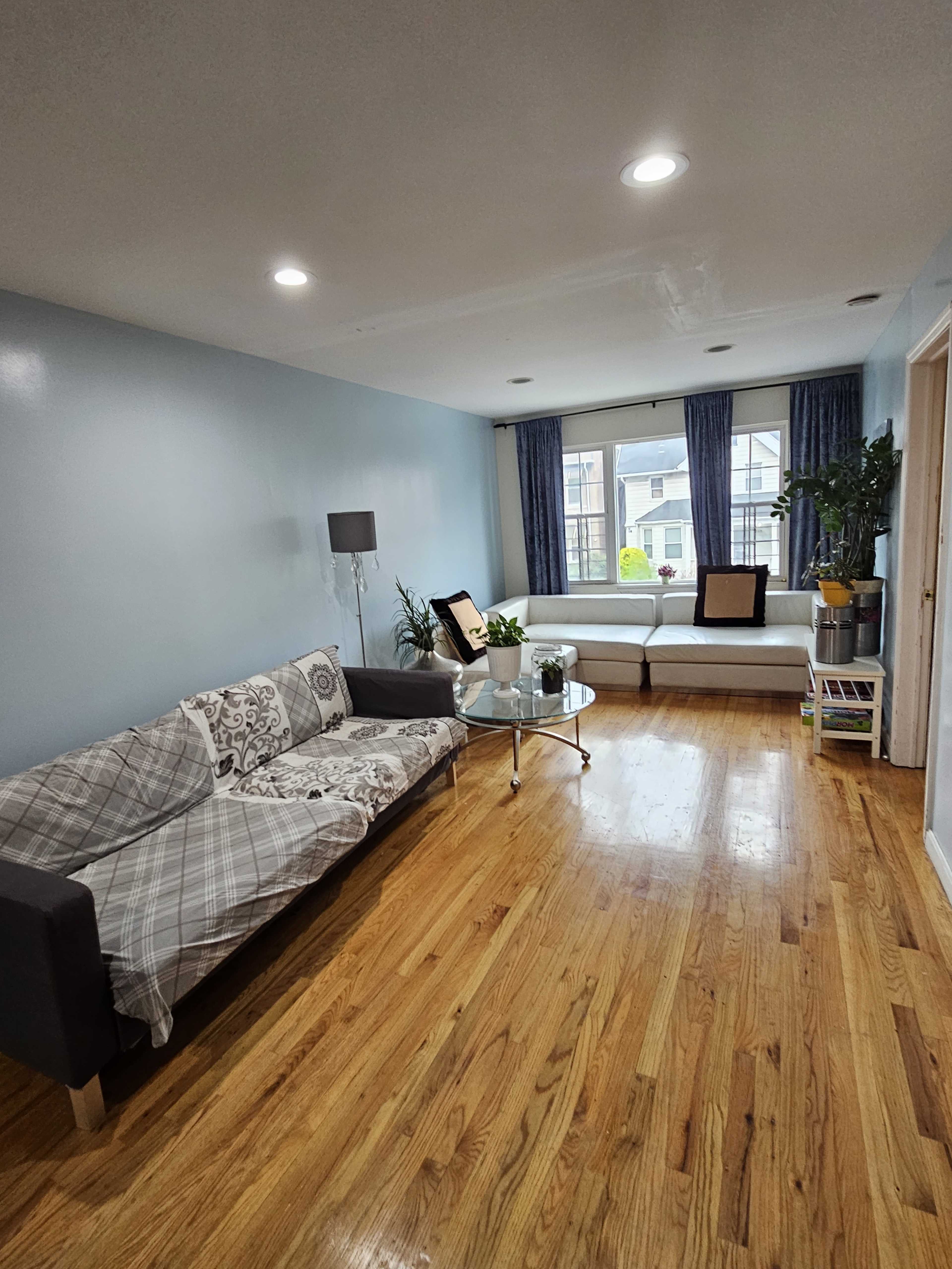 A living room featuring a couch with a patterned throw, a glass coffee table, and large windows with blue curtains.