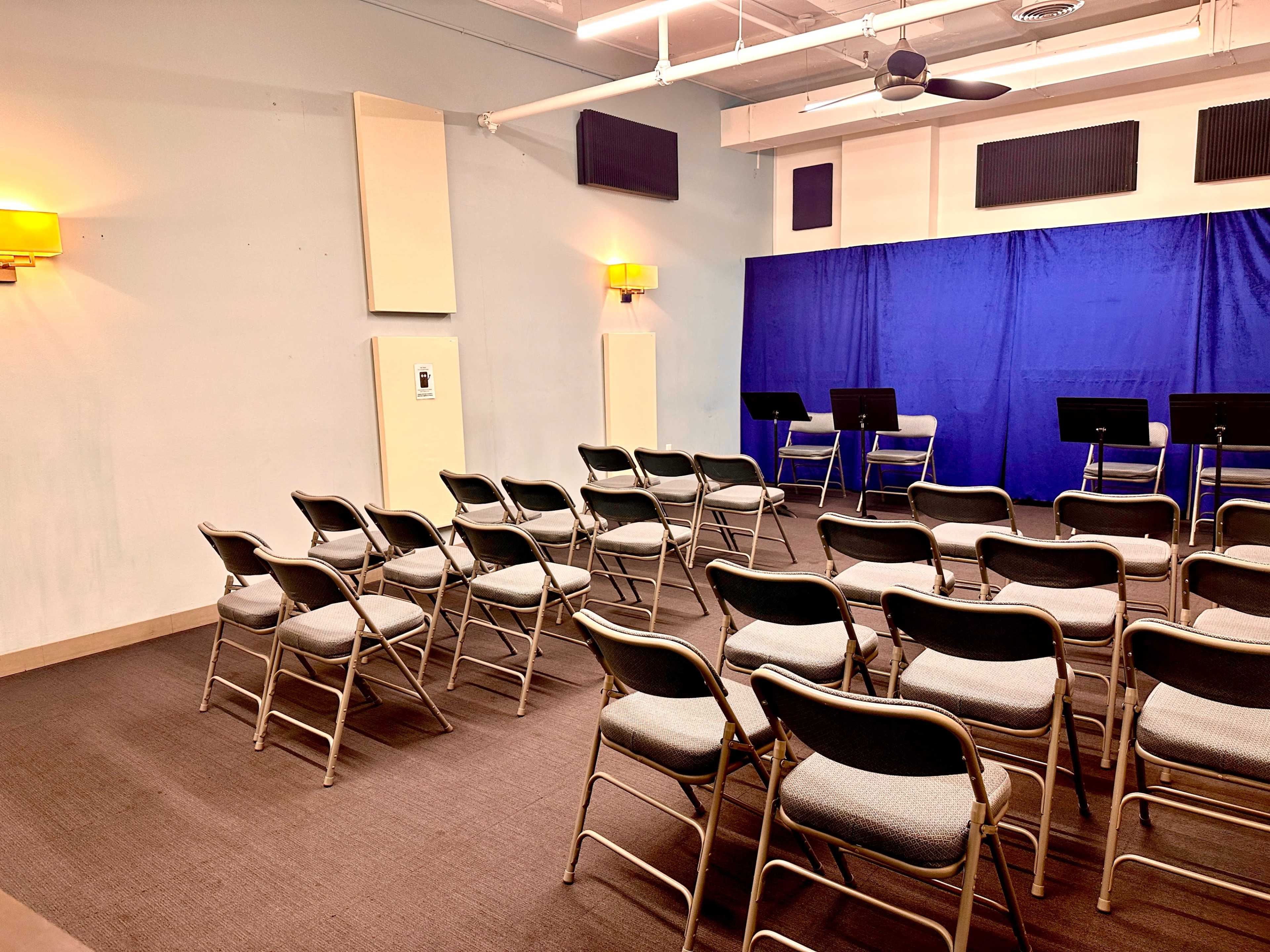 The image shows a small room with rows of folding chairs facing a blue curtain and two music stands.