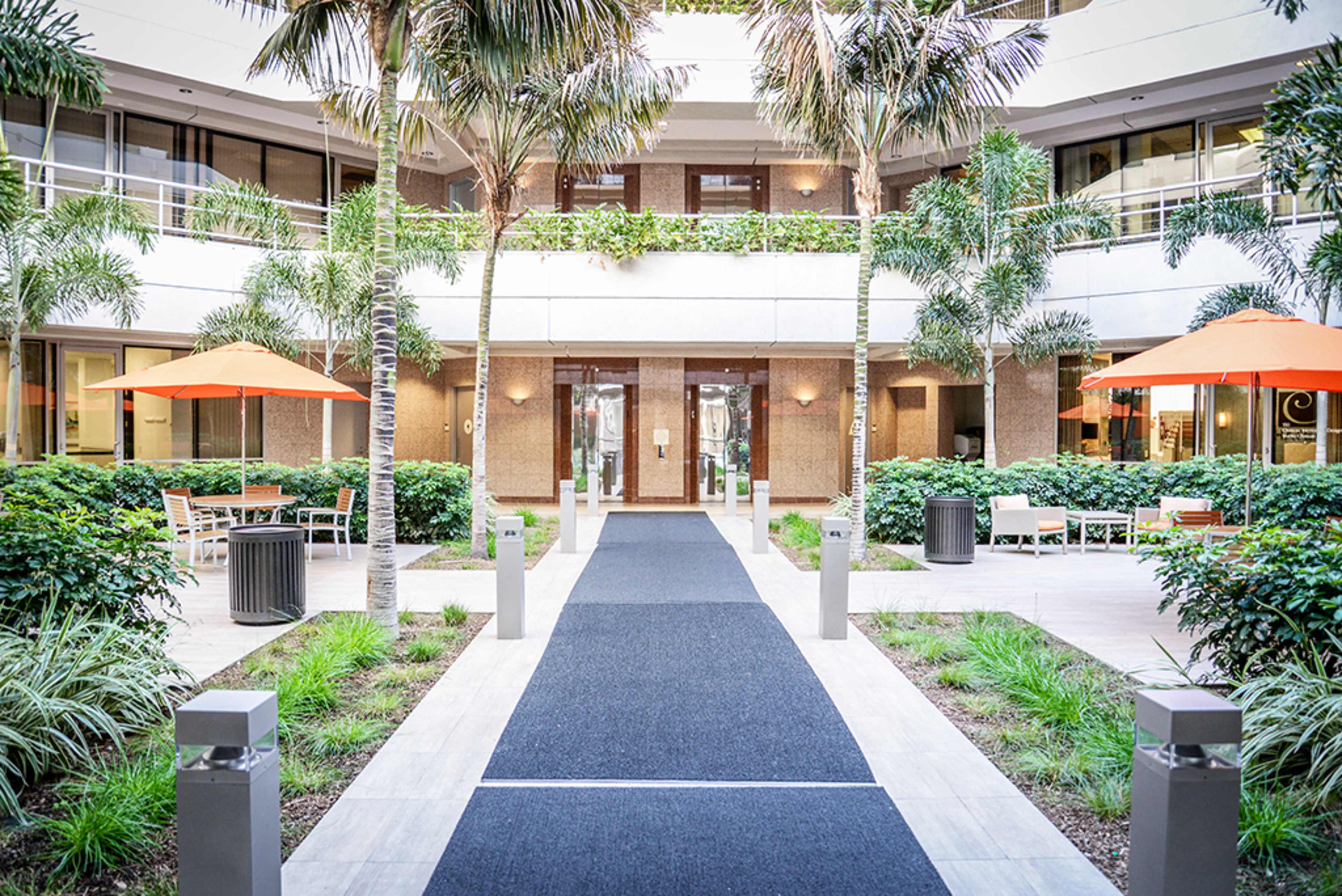 A tropical courtyard with palm trees, orange umbrellas, and a central pathway leading to a glass entrance.