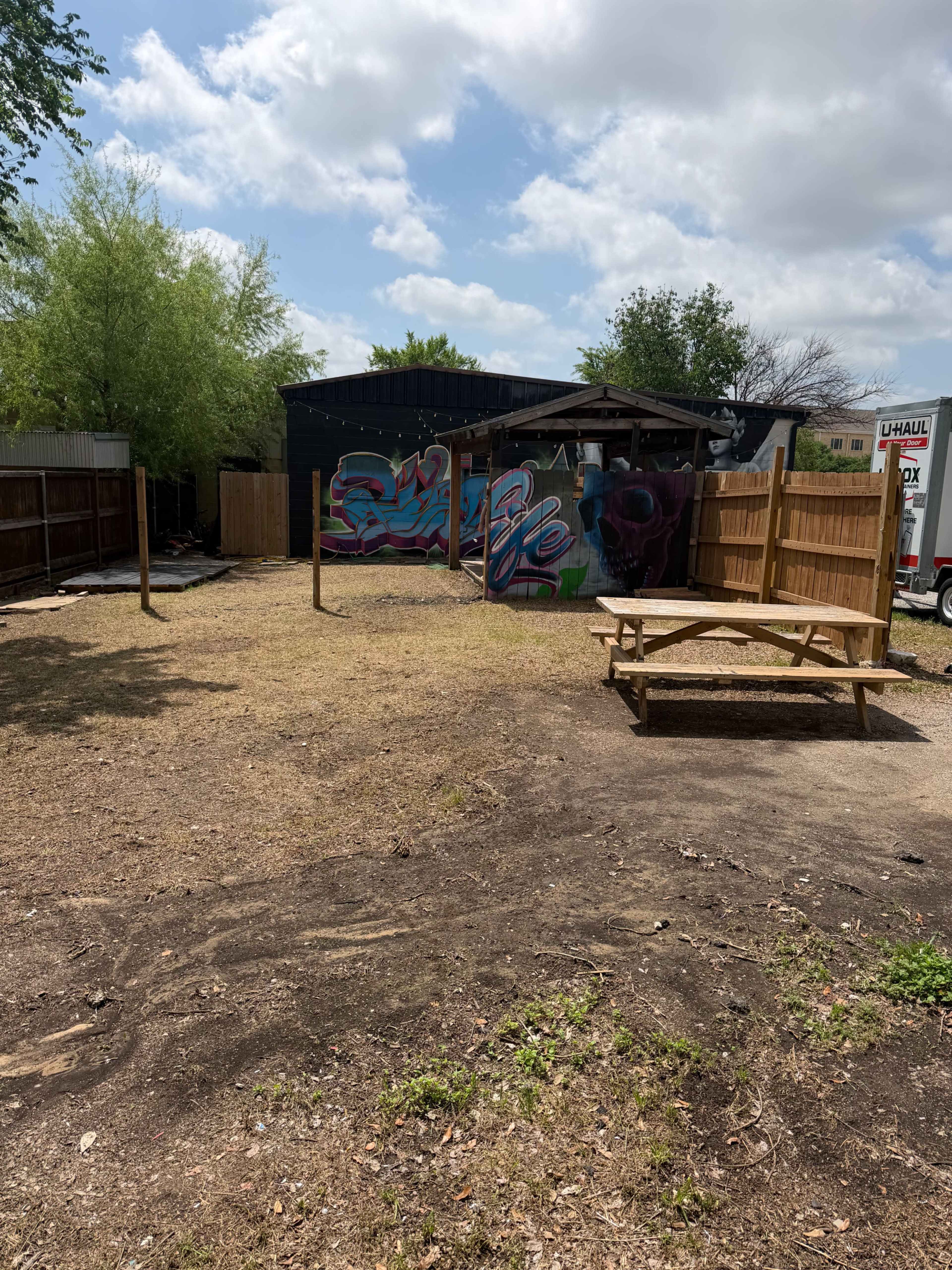 The image shows an outdoor area with a wooden picnic table, a fenced space, and a colorful mural on a wall in the background.