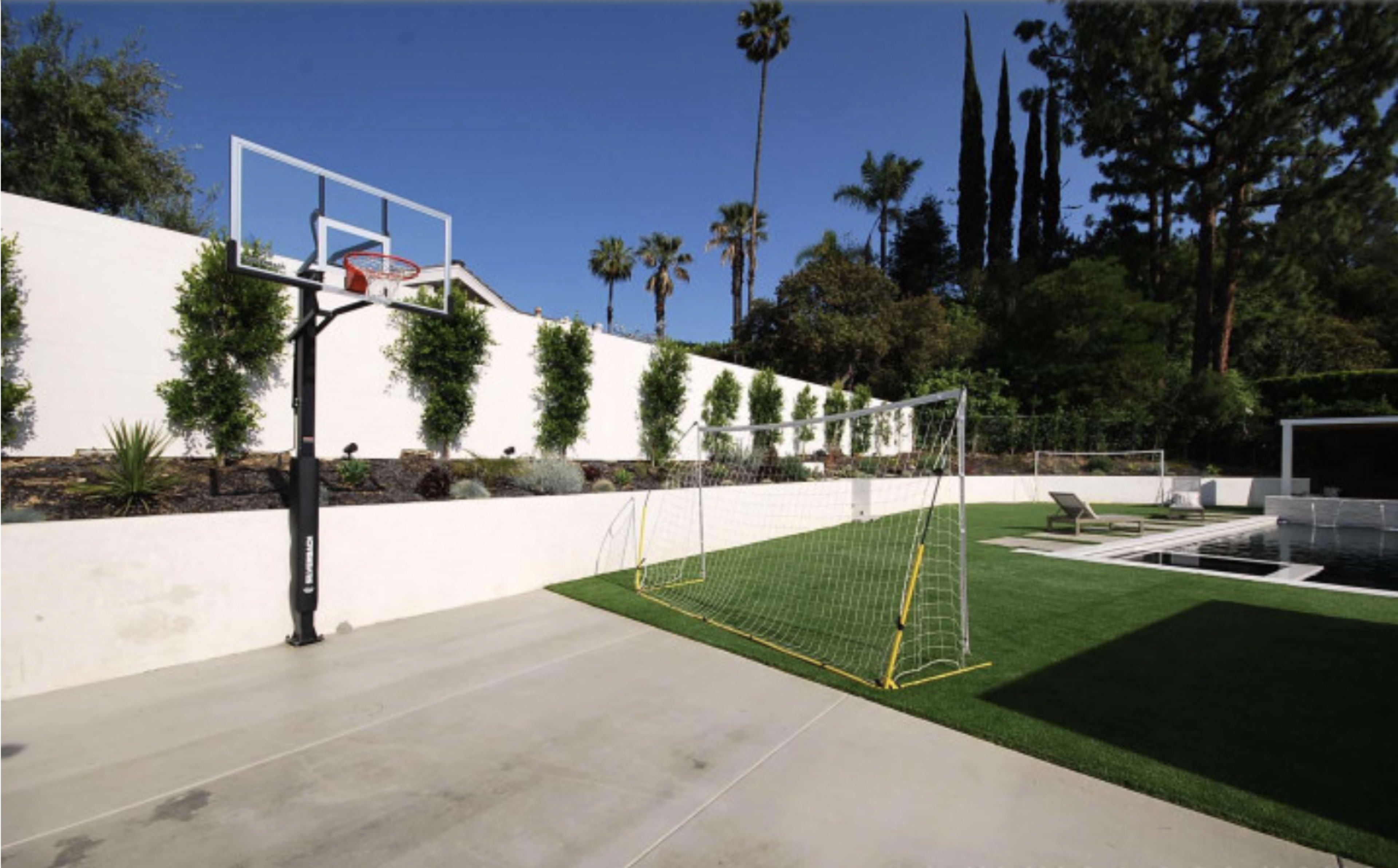 A backyard featuring a basketball hoop, two soccer goals, and a swimming pool surrounded by grass and palm trees.