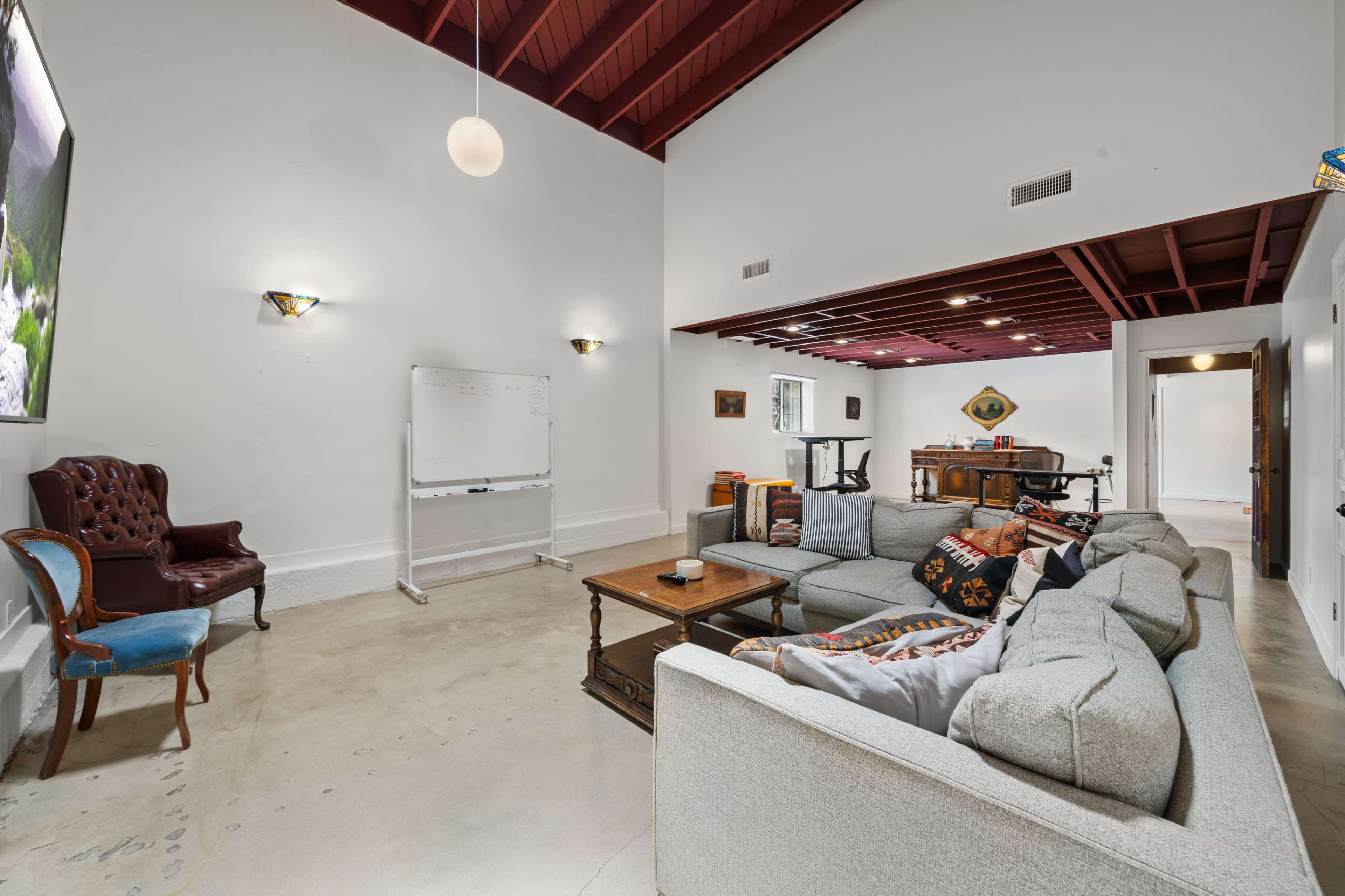 A spacious living room features a large sectional sofa, a coffee table, a flat-screen TV, and a whiteboard, with a dining area visible in the background.