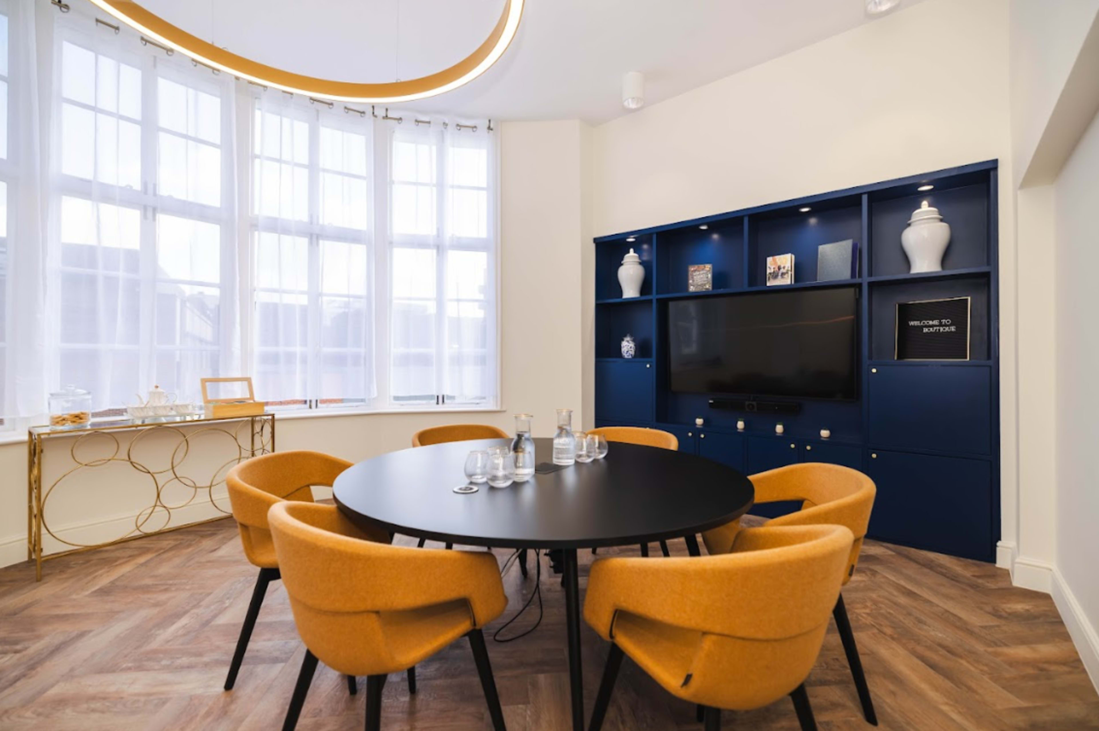 A modern meeting room with a round black table surrounded by orange chairs, located near large windows and a blue shelving unit with decorative items.