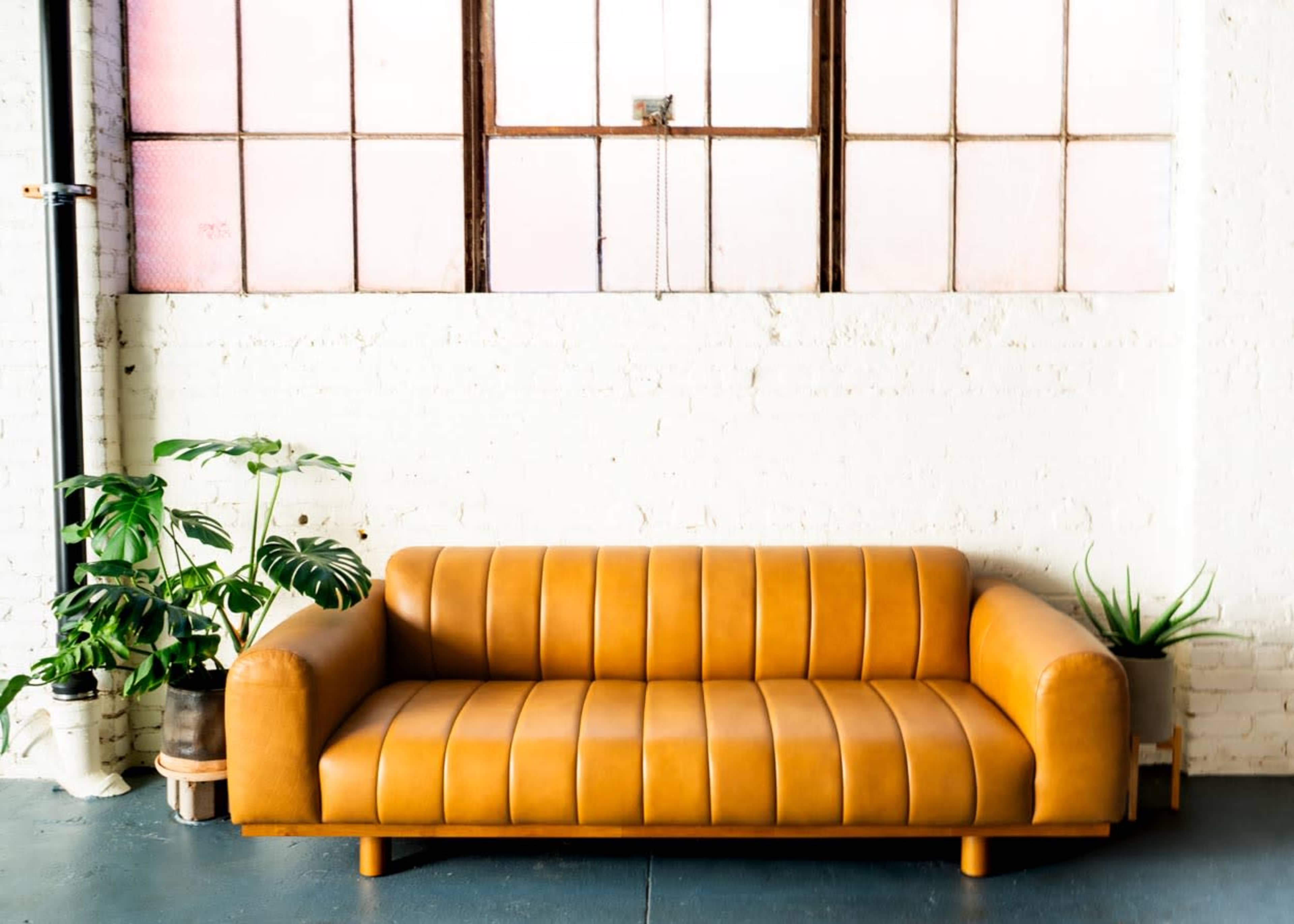 A brown leather sofa is positioned against a white brick wall beneath large windows with pink-tinted glass.