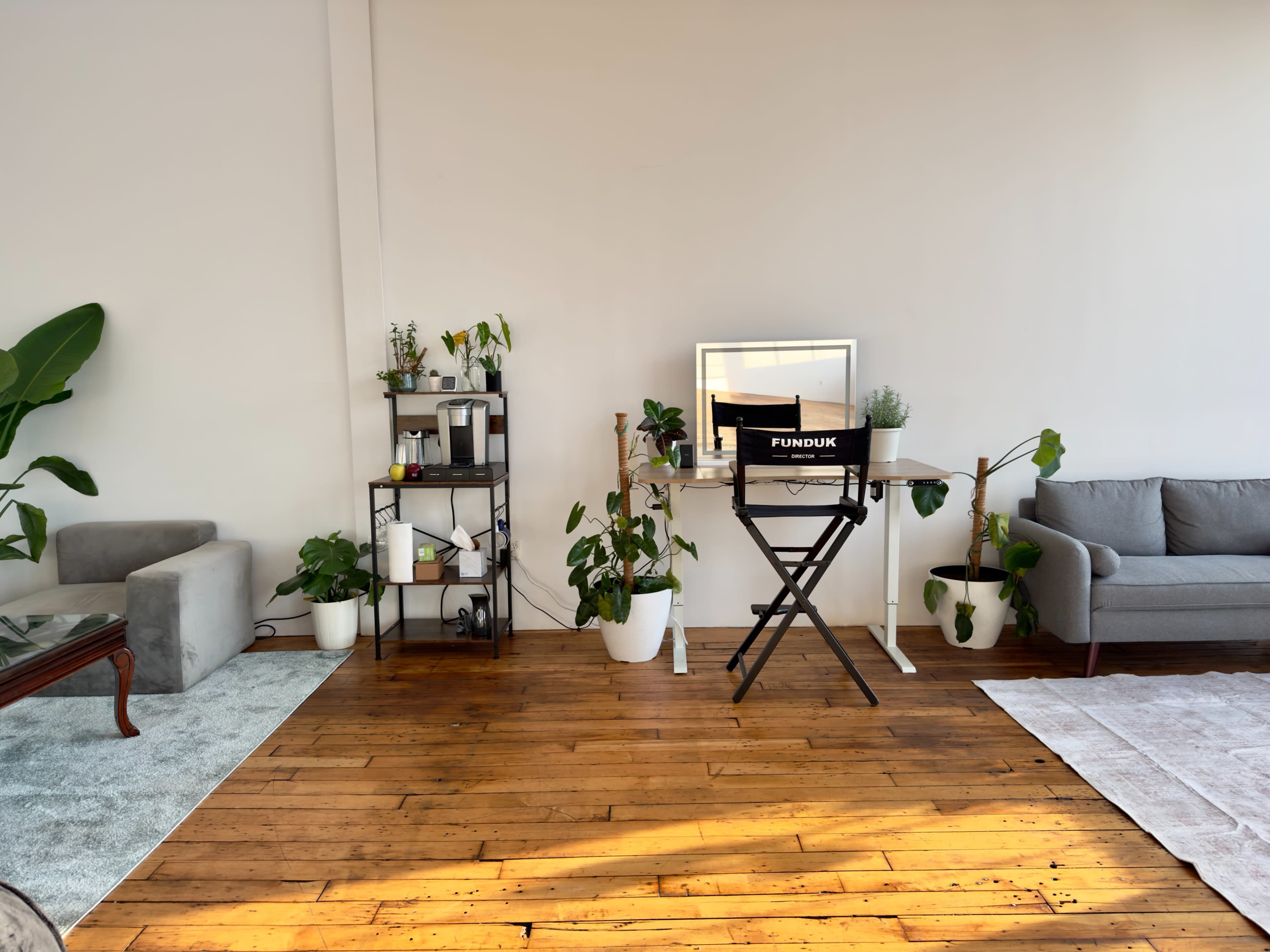The image shows a bright room with wooden floors, featuring a gray couch, a shelving unit with plants, a mirror, and a director's chair labeled "FUNDUNK."
