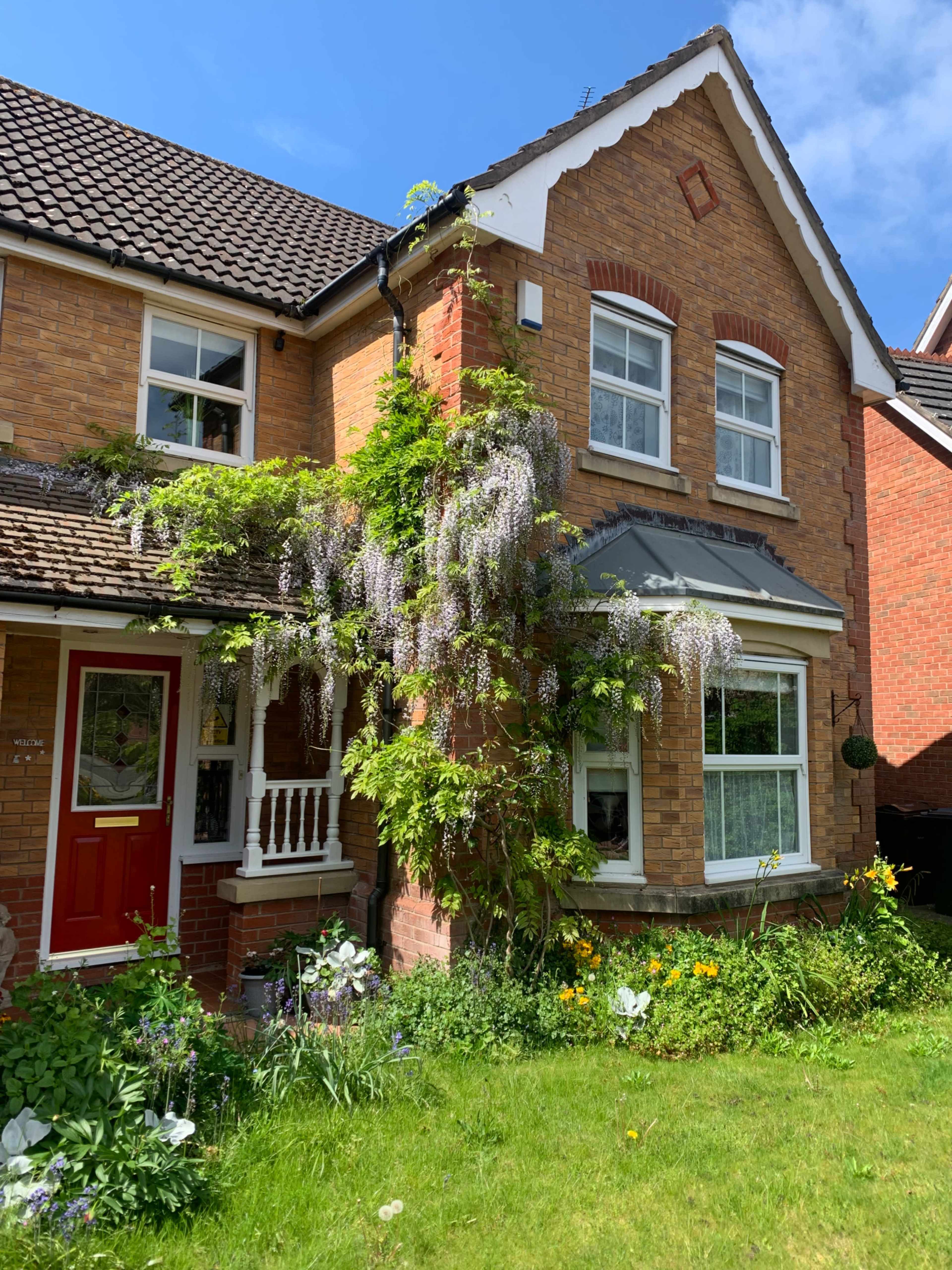 A two-story brick house features a red front door and is adorned with climbing wisteria and various flowers in the garden.