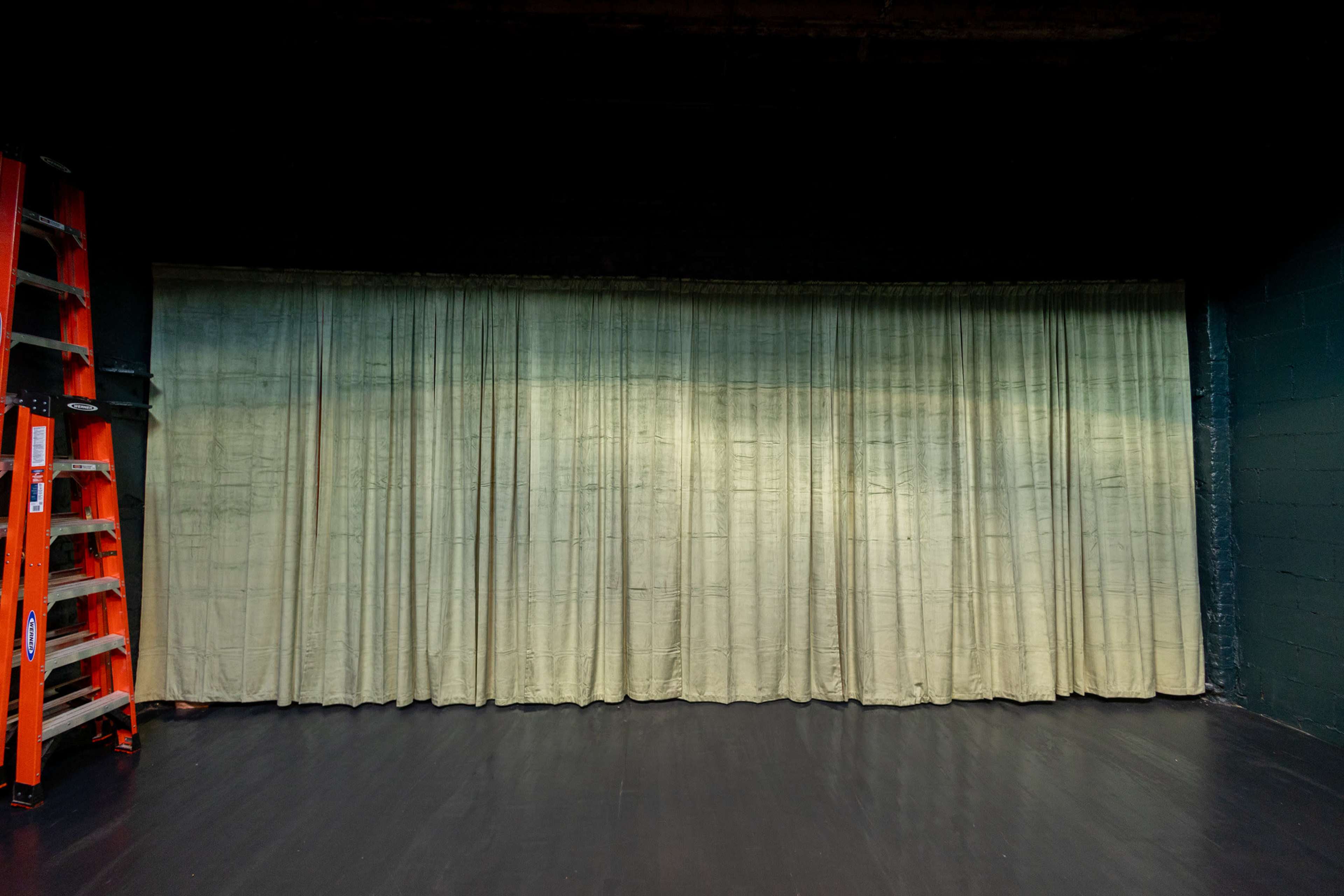 A green curtain hangs on a stage wall near an orange ladder.