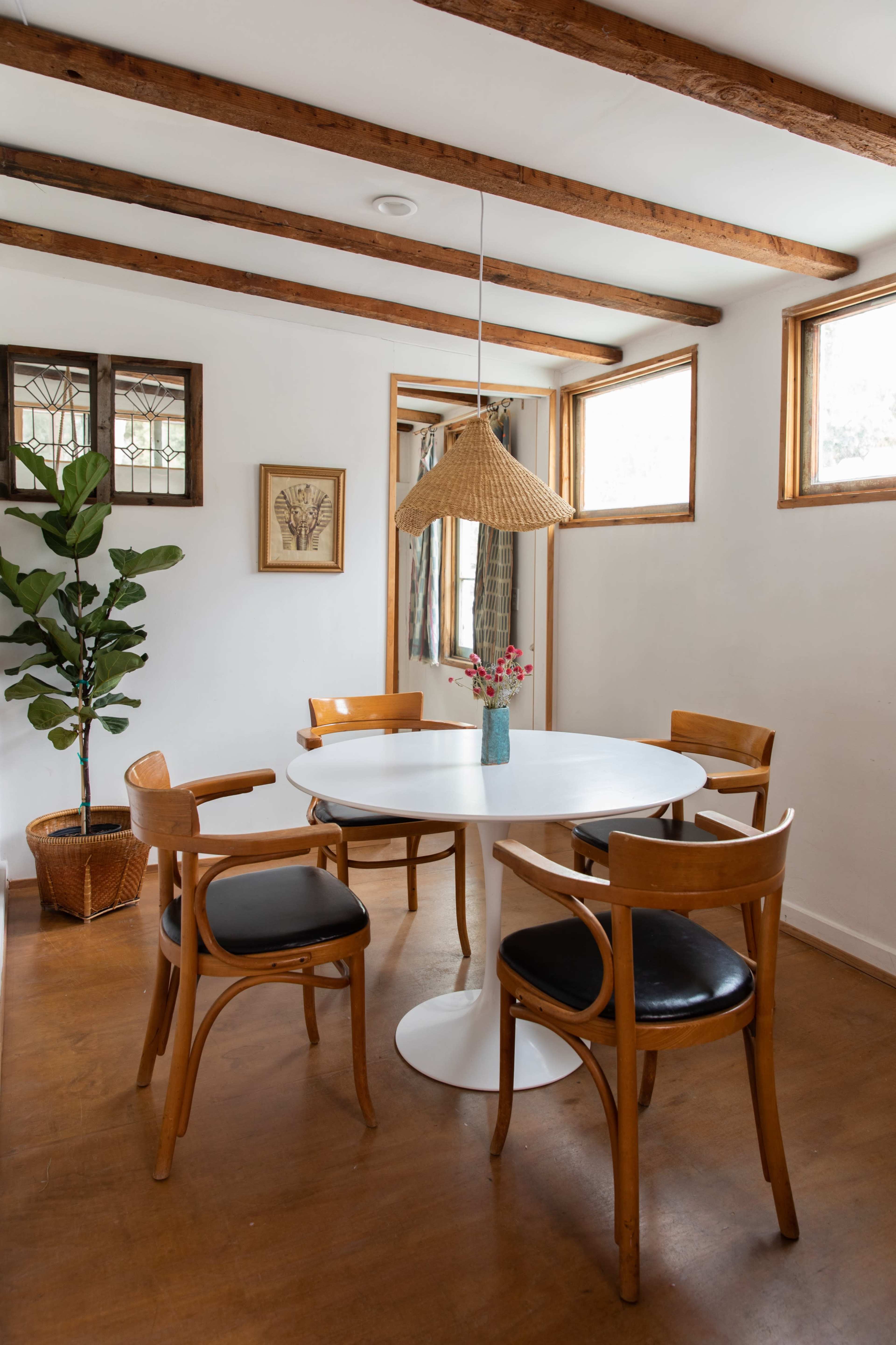 The image shows a dining area with a round white table surrounded by four wooden chairs, a plant in a basket, and natural light coming through the windows.
