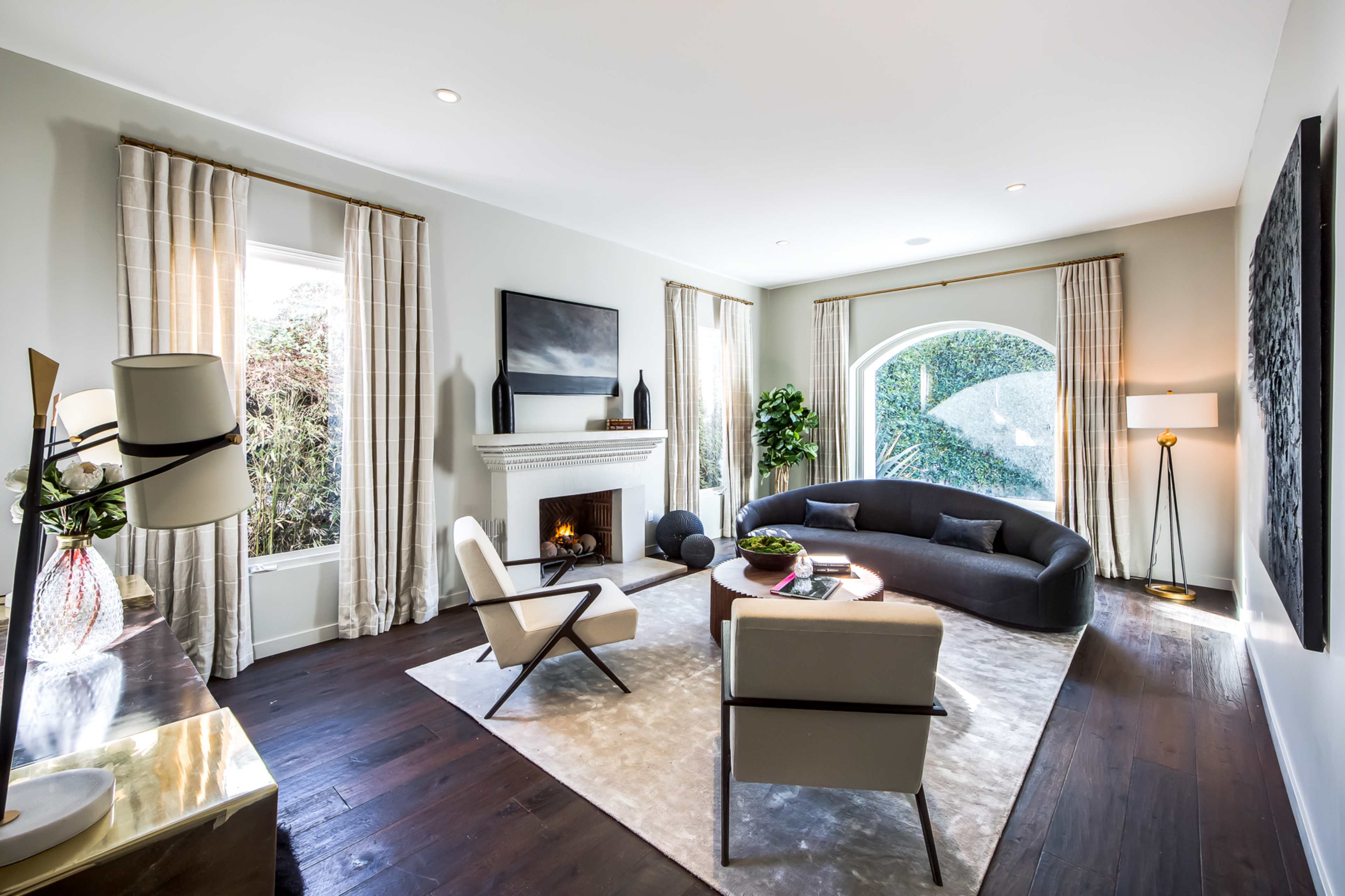 The image shows a modern living room featuring a curved sofa, two armchairs, a fireplace, and large windows draped with light curtains.