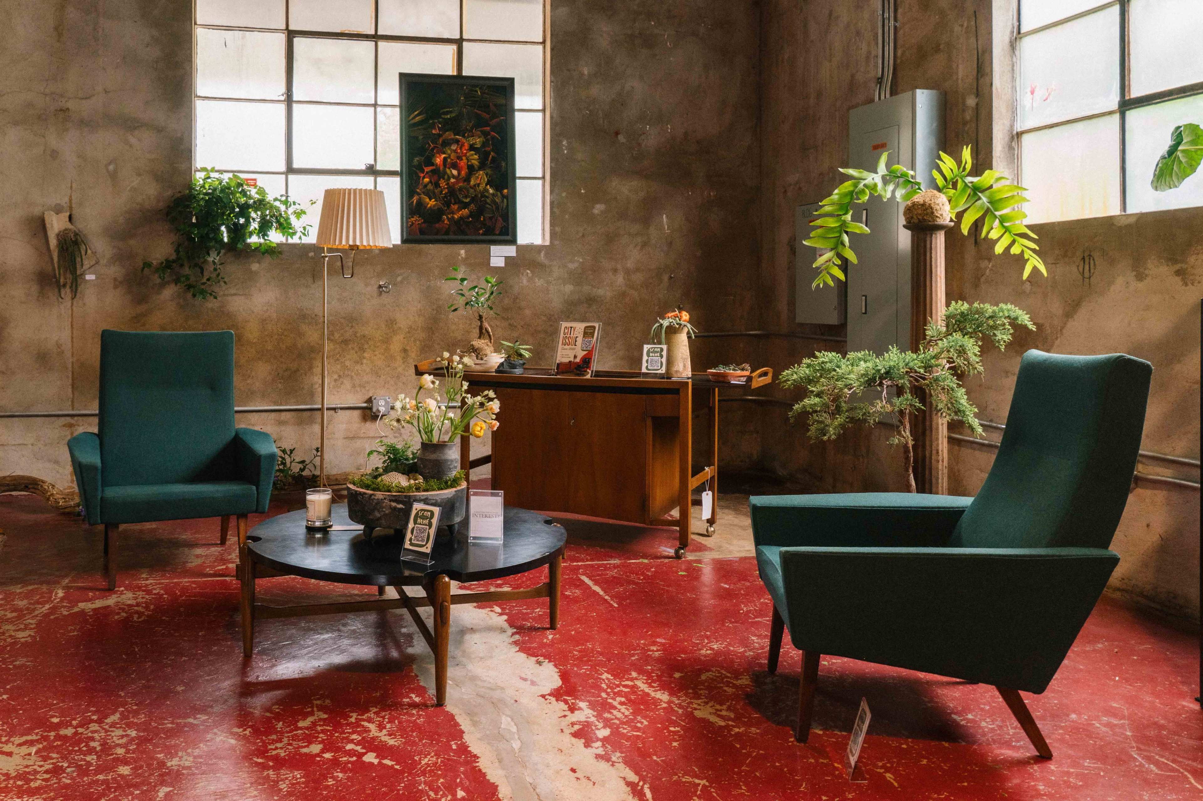 The image shows a vintage-inspired living space with two green chairs, a round table, and a wooden desk, against a backdrop of peeling walls and large windows.