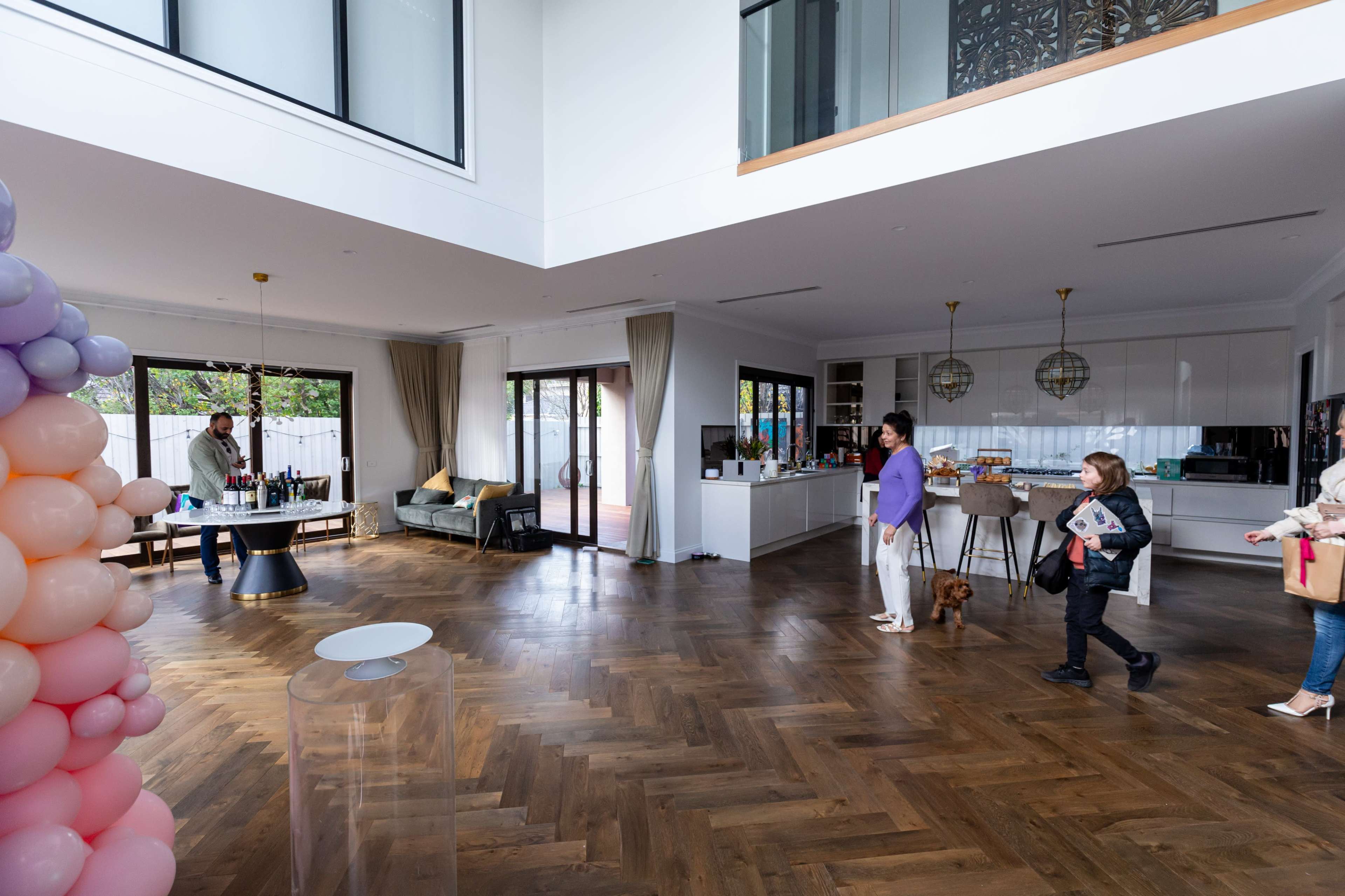 A spacious modern interior with wooden flooring, a balloon arch, guests mingling, and a bar area in the background.