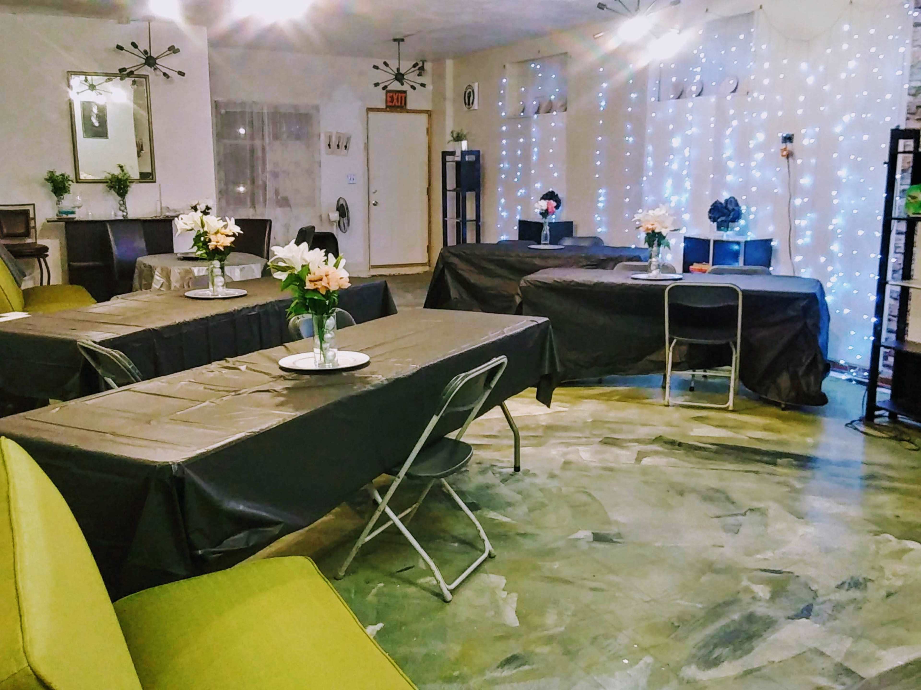 The image depicts a room set up for an event, featuring several tables covered with black tablecloths and floral centerpieces, with soft lighting and decorative fairy lights in the background.