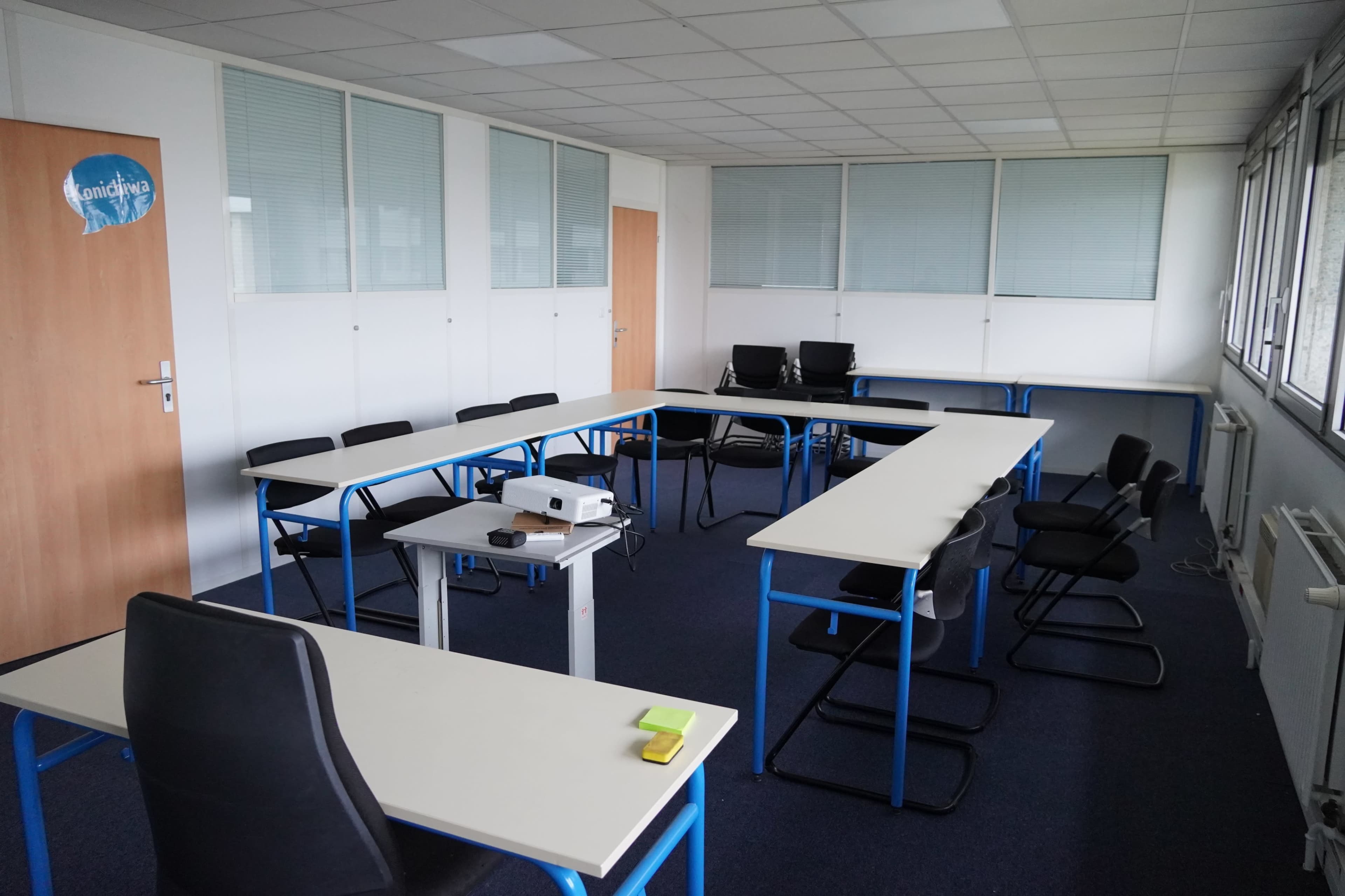 A bright, empty classroom features a large U-shaped arrangement of tables and chairs, with a projector set up on a small table.
