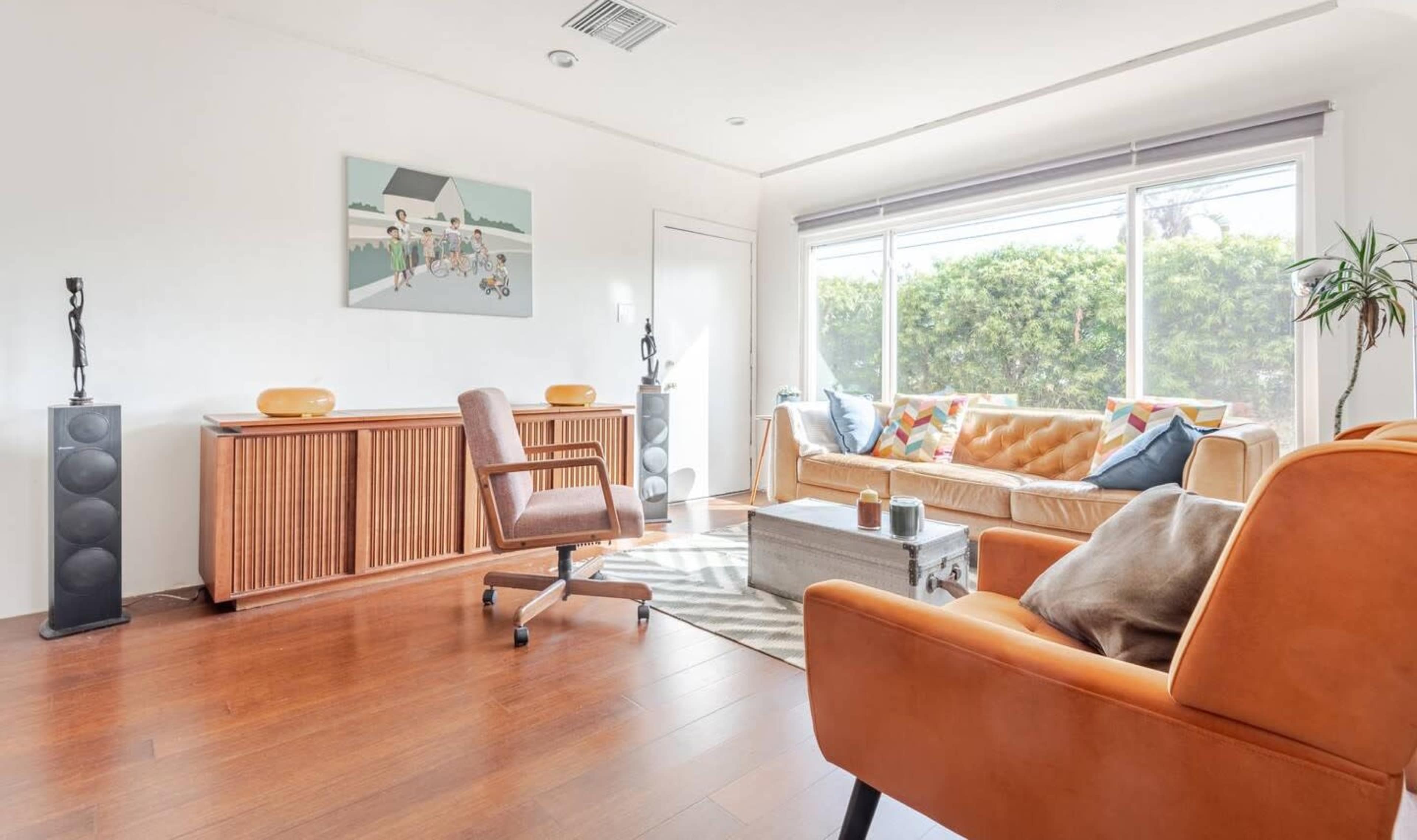 A well-lit living room features a sofa, two chairs, a wooden cabinet, and large windows with a view of greenery outside.