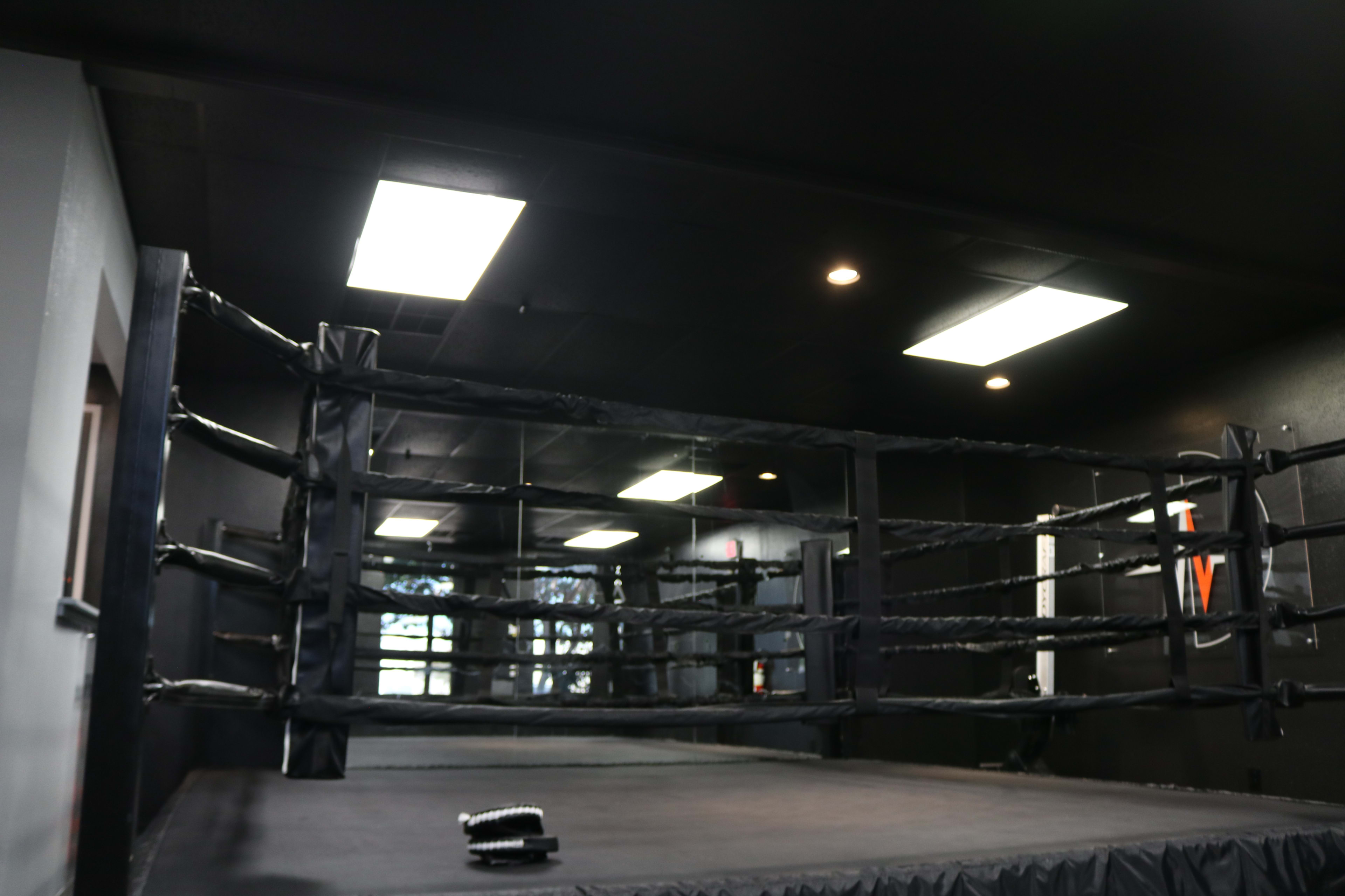 An empty boxing ring situated in a dimly lit gym with black walls and overhead fluorescent lights.