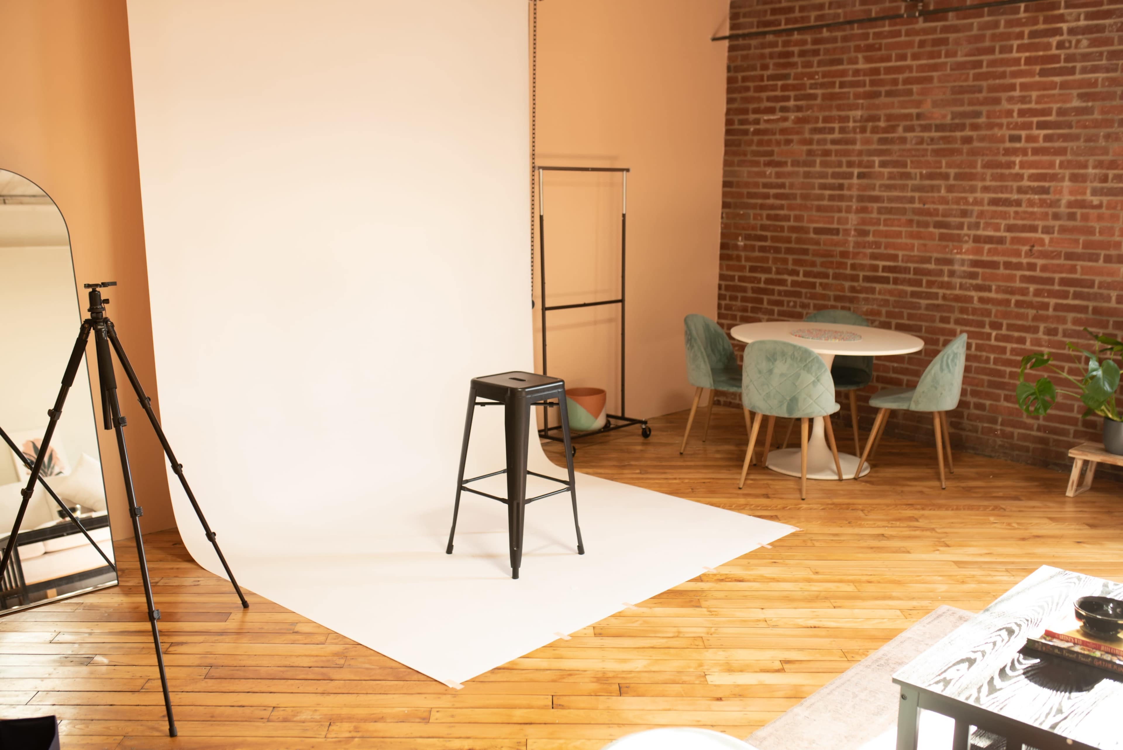 A studio space with a black stool on a white backdrop, surrounded by a wood floor, a round table with chairs, and a tall tripod.