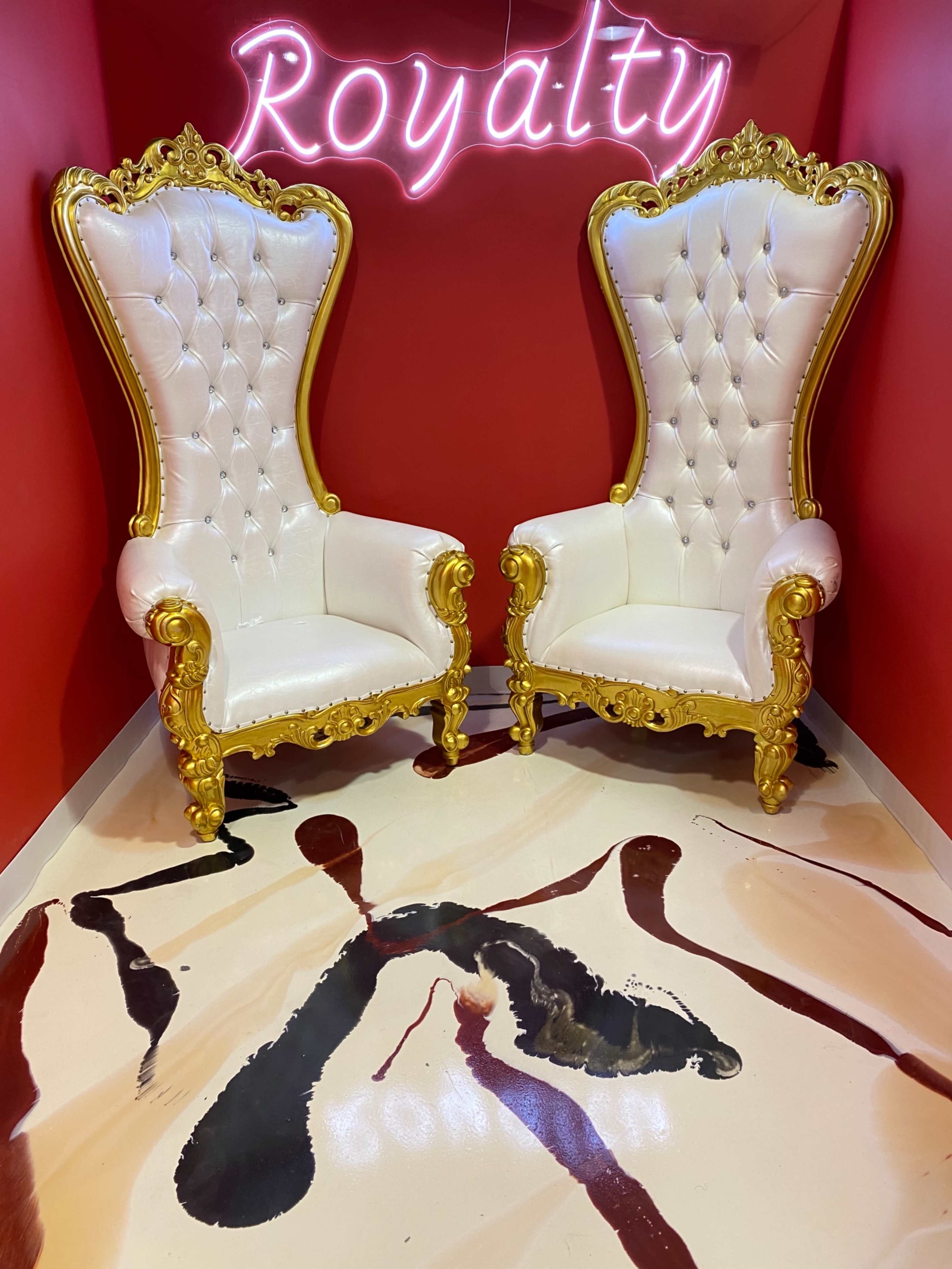 Two ornate, white and gold throne chairs positioned on a floor with a paint-like pattern, beneath a neon sign that reads "Royalty."