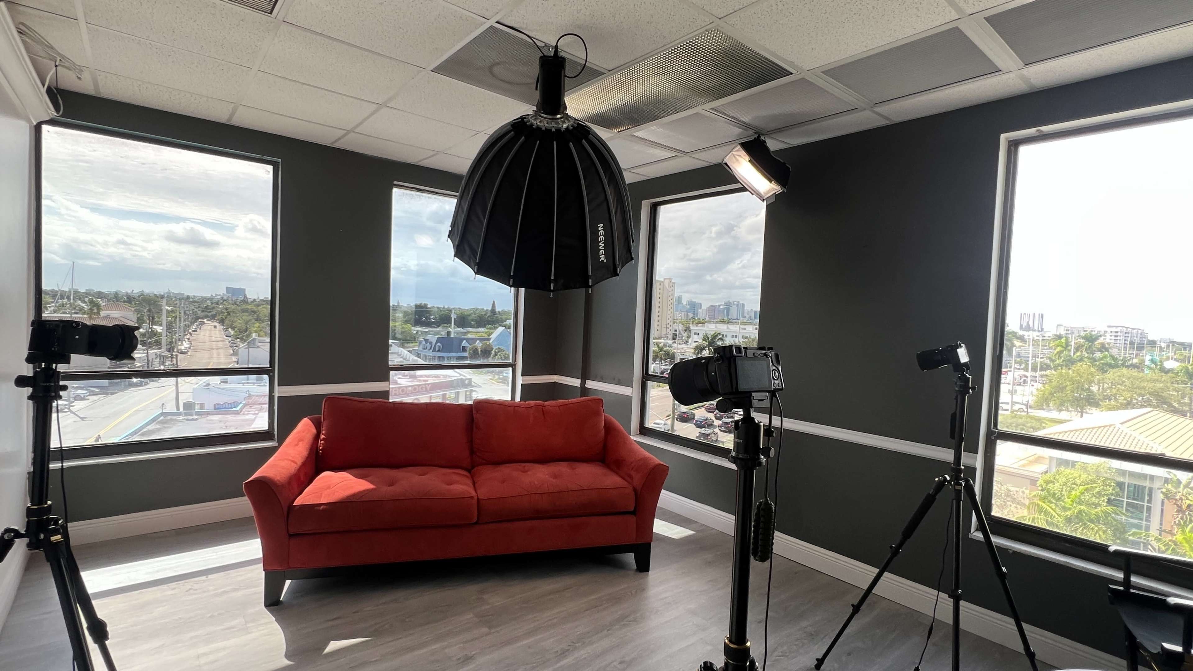 The image shows a brightly lit studio space featuring a red sofa, two camera lights, and large windows offering a view of the city outside.