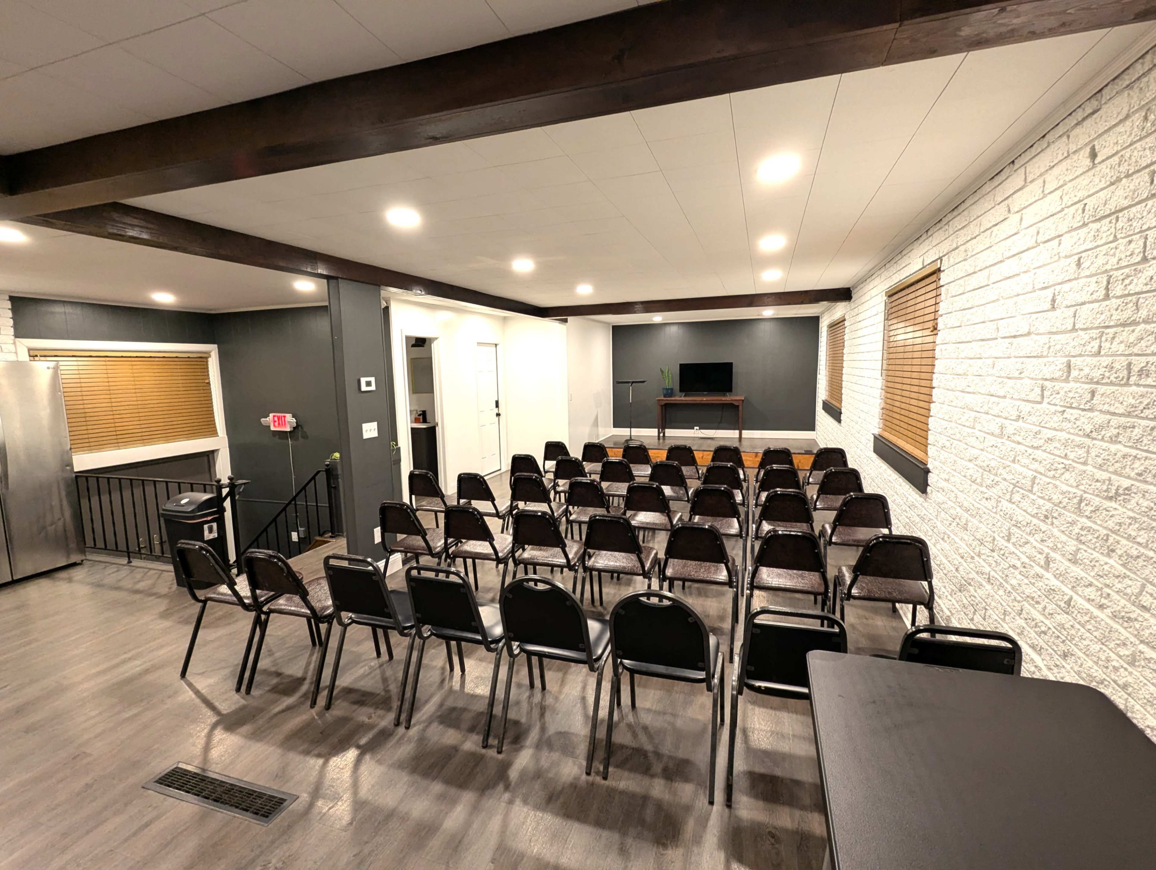 The image shows a room with a row of black chairs arranged facing a small stage or presentation area, with a flat screen monitor and a table at the front.