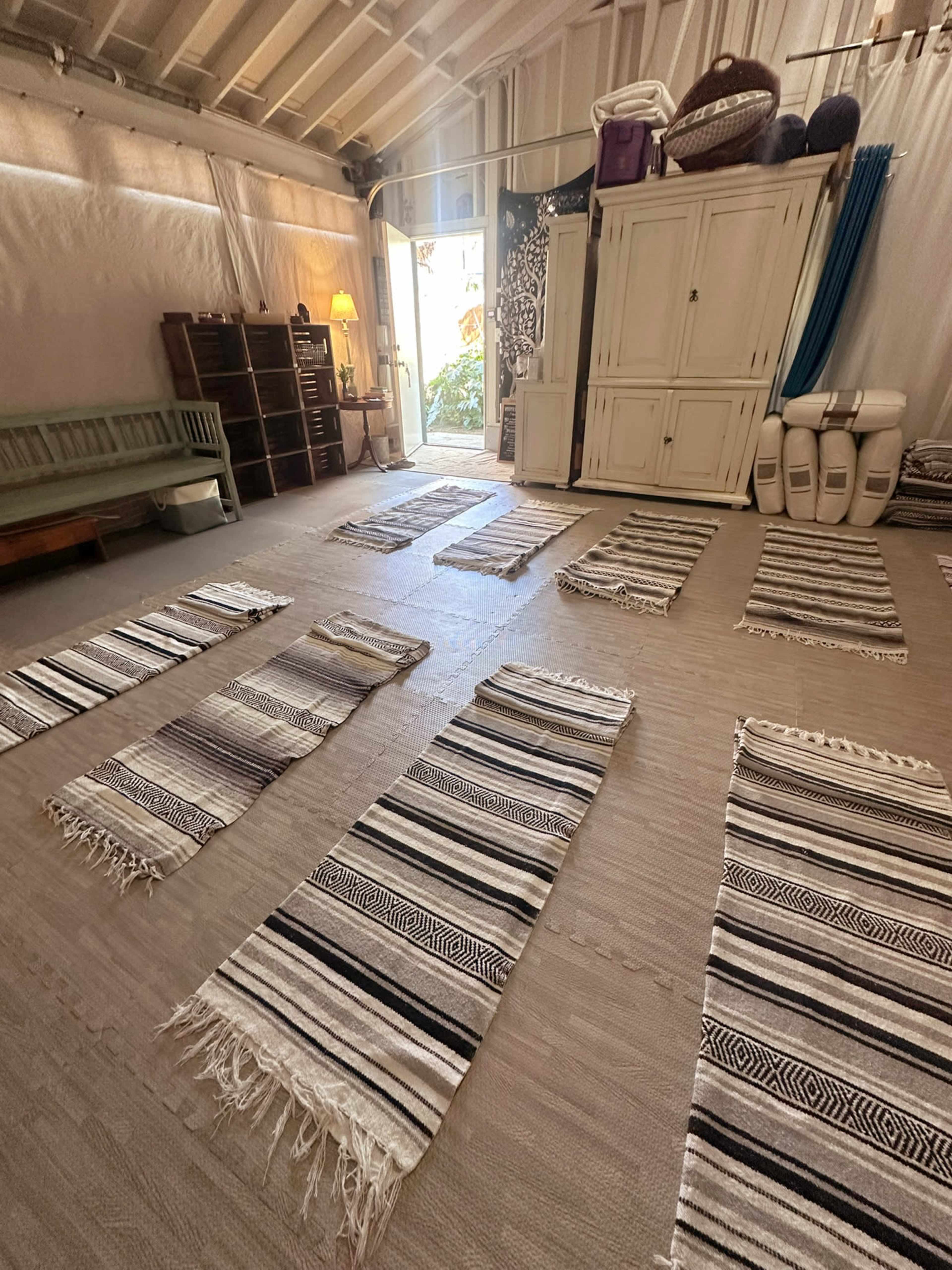 A spacious room features several woven rugs arranged on the floor, with a doorway leading to an outdoor area visible in the background.