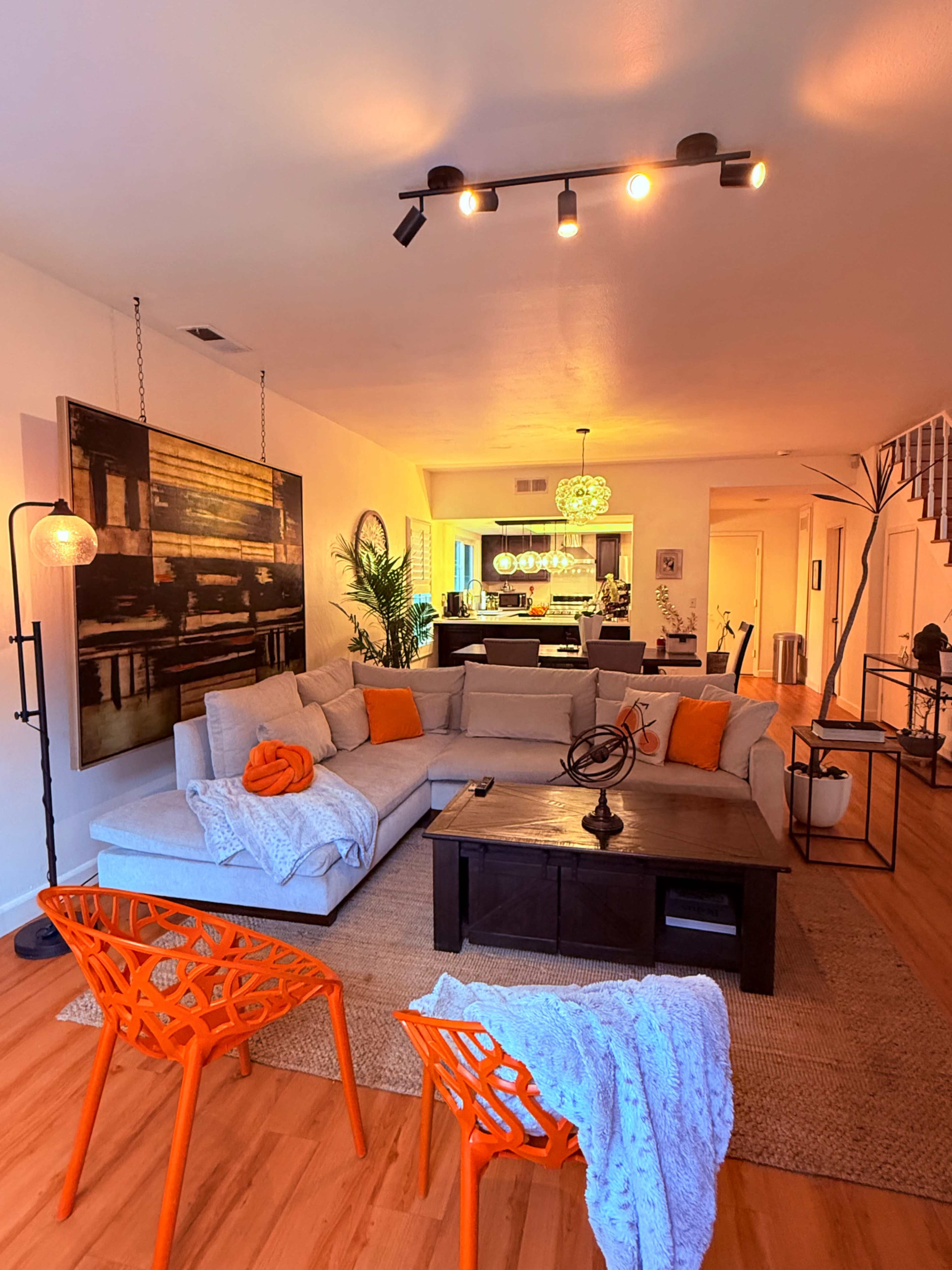 A modern living room features a gray sectional sofa with orange accents, an illuminated bar area in the background, and orange chairs arranged around a coffee table.