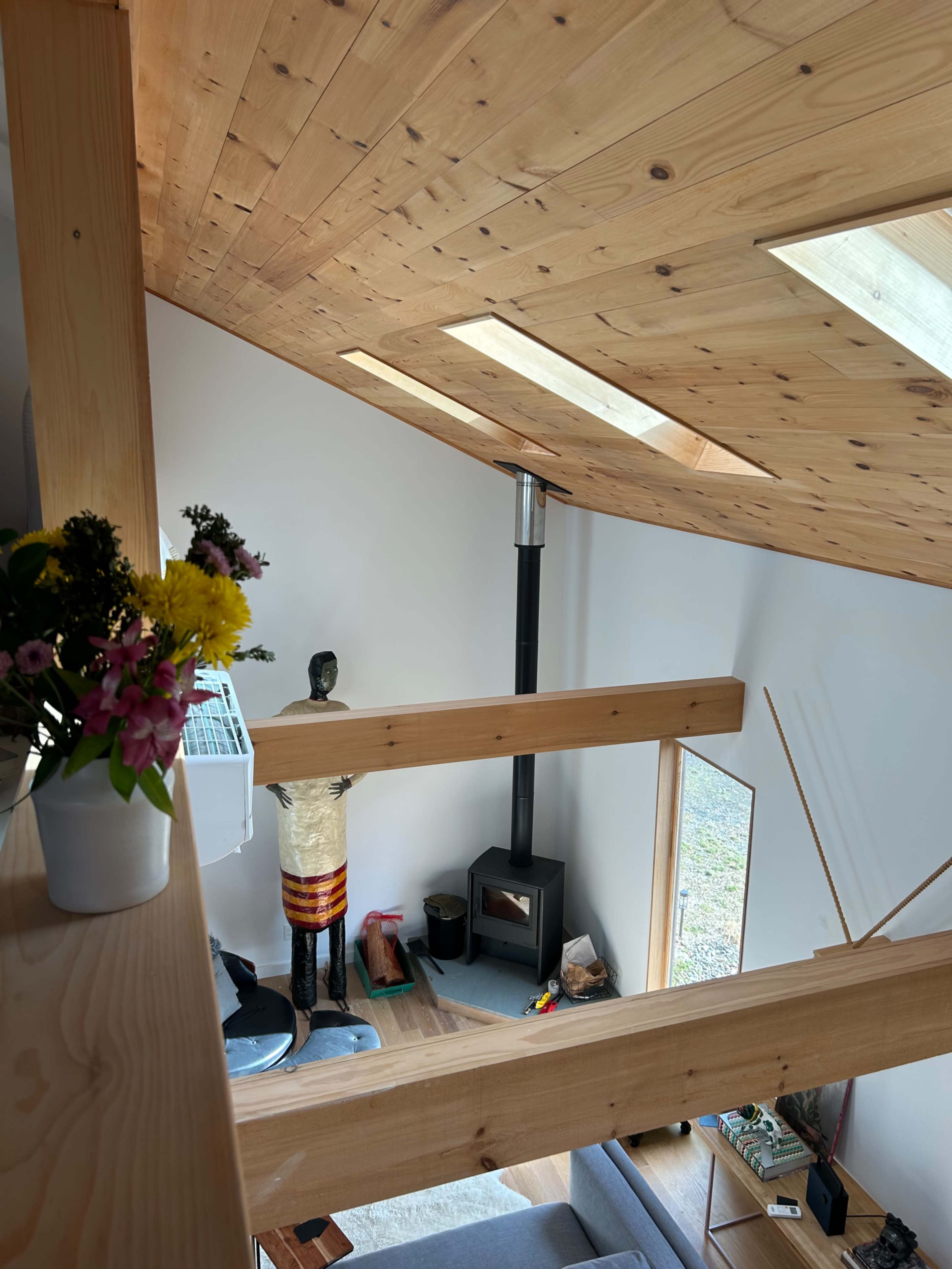 The interior of a modern wooden cabin with a sloped ceiling, featuring a wood stove and a balcony view of the living space below.