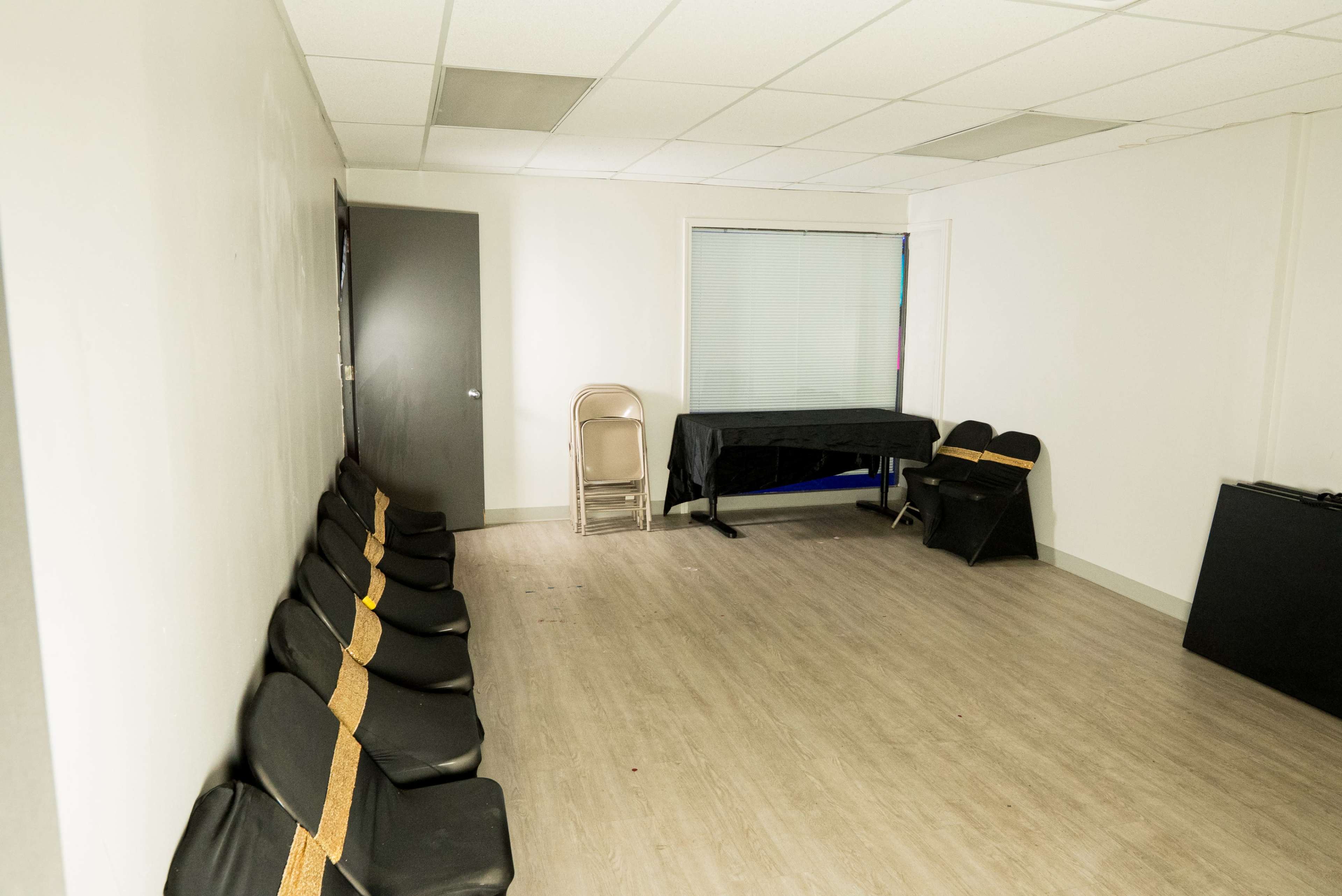 The image shows a sparsely furnished room with several black chairs arranged in a line, a folding chair stacked nearby, a table covered with a black cloth, and a window with blinds.
