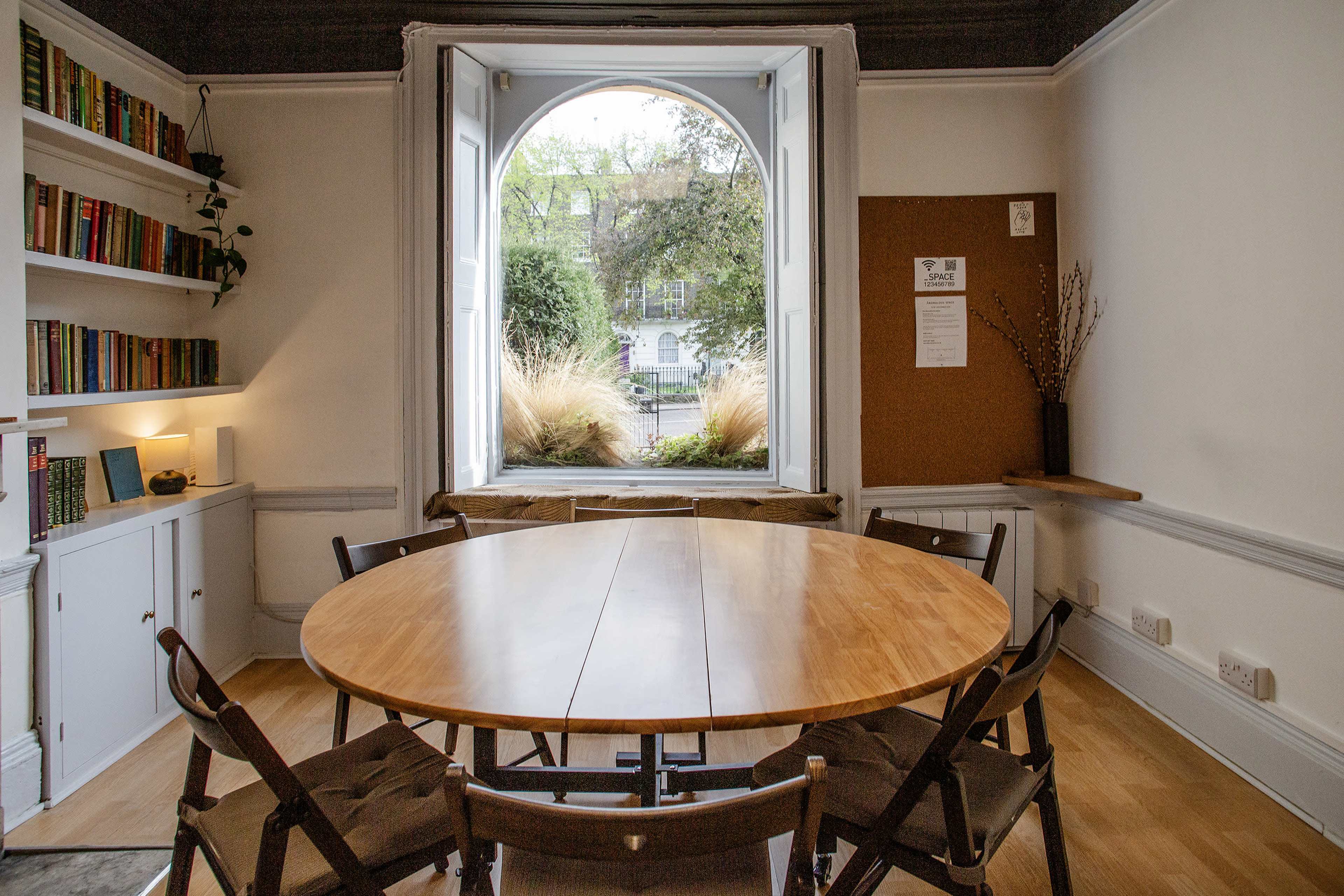 A round wooden table surrounded by chairs is positioned in a well-lit room with a large circular window overlooking a garden.
