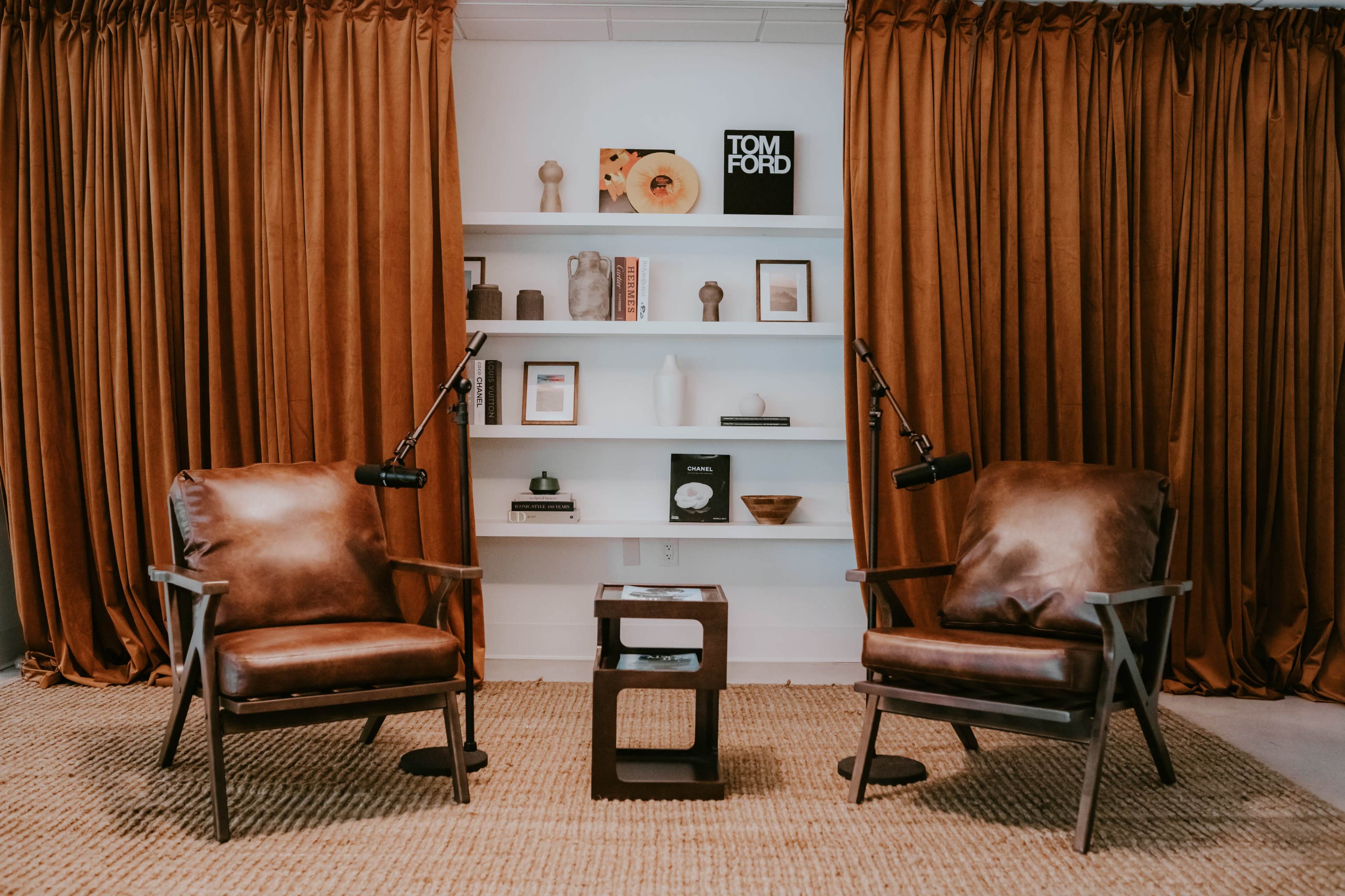 The scene features two brown leather chairs facing a small dark table, set against a backdrop of orange curtains and a bookshelf with decorative items.
